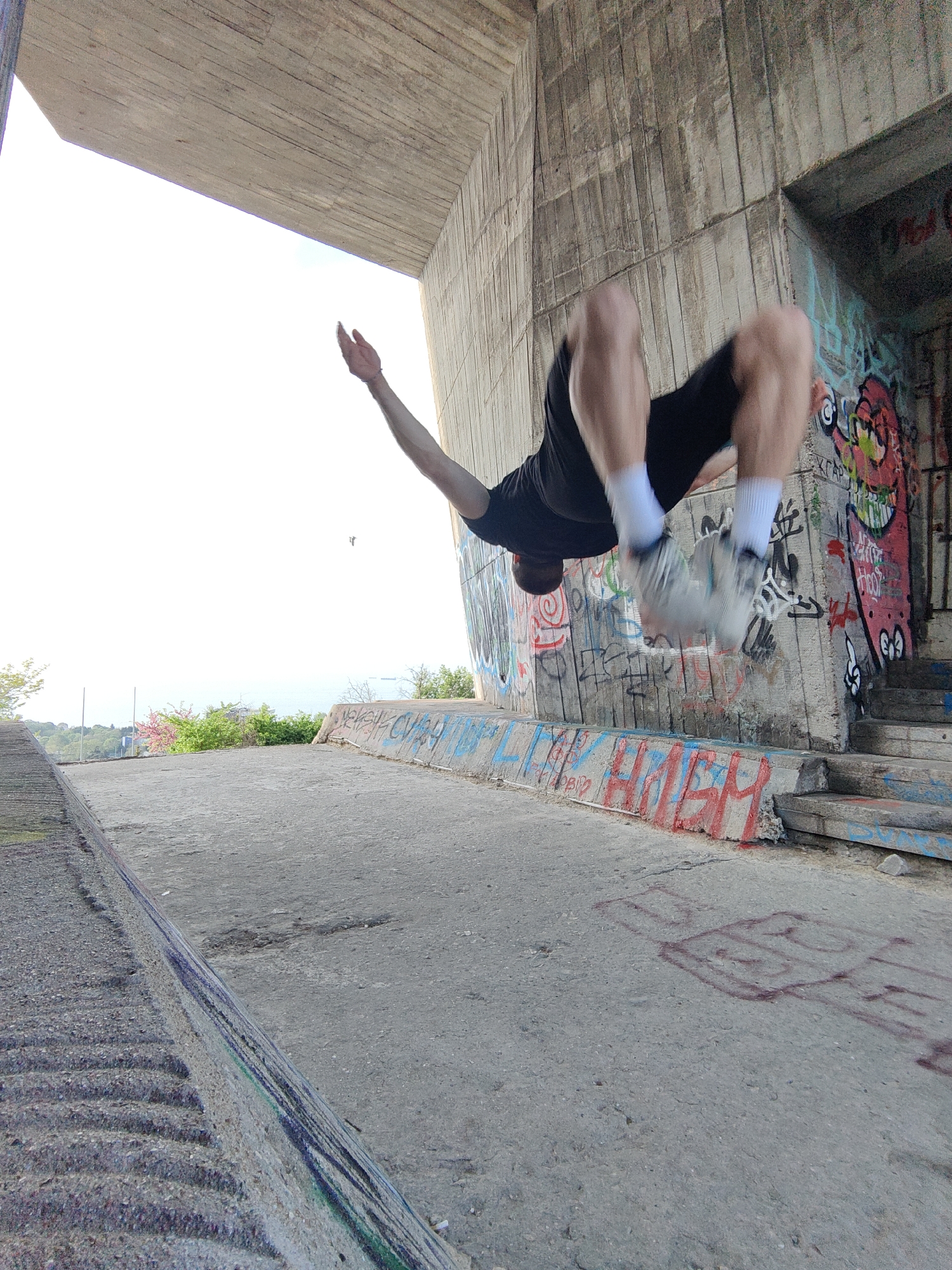 A man performs a backflip under a concrete structure. The man is wearing a black t-shirt, black shorts, white socks, and light-colored athletic shoes. His left arm is extended upwards. The background features a concrete wall covered in graffiti, stairs, a concrete ledge with graffiti, and greenery in the distance. The location is Varna, Bulgaria.