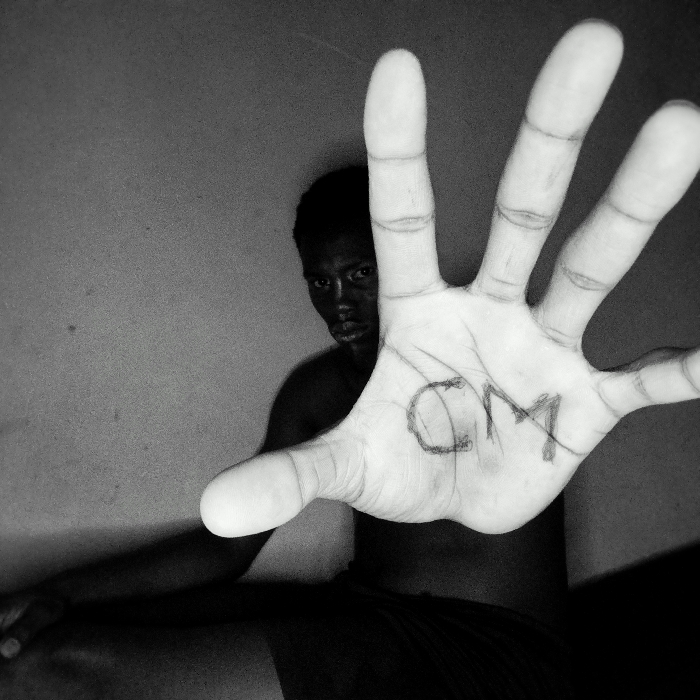 A monochrome photograph features a dark-skinned person behind a dramatically oversized hand. The hand, extended toward the viewer, dominates the frame. "CM" is crudely written on the palm. The person's face is partly obscured, but their expression appears serious. The background is a plain, slightly textured wall. The lighting emphasizes the contrast between the hand and the individual, creating a sense of depth and perspective distortion.