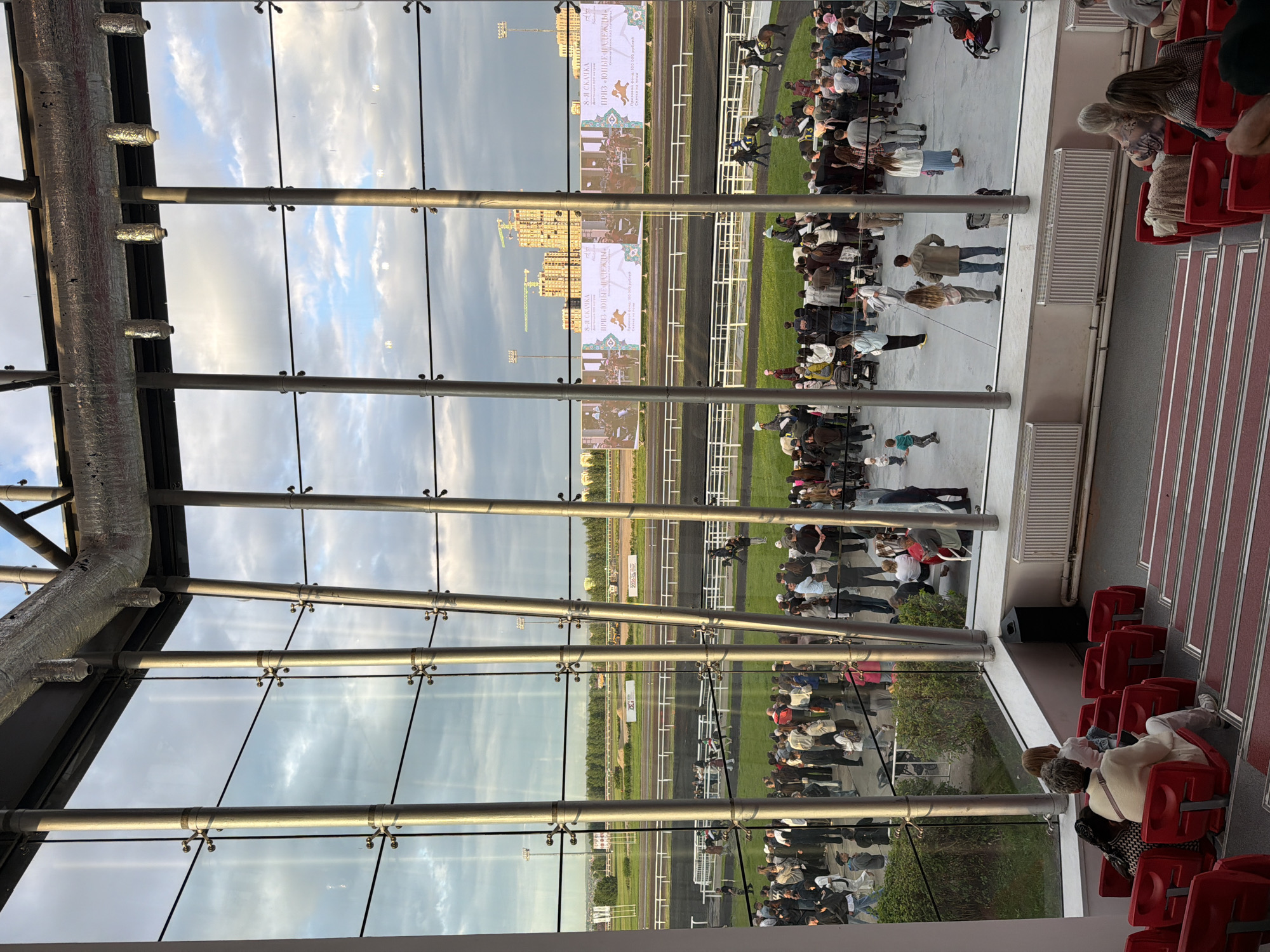The image shows a reflection of a horse race on a large glass structure. The reflection captures the race track, spectators in the stands, and large video screens displaying race information. The spectators are dressed in casual attire, suggesting an outdoor event. The weather appears to be partly cloudy, with the sky visible in the reflection. The large glass panes are supported by metal beams, indicating a modern architectural design. The screens display text in Russian, identifying the event as "8-Я СКАЧКА ПРИЗ «ЮНЫЕ НАДЕЖДЫ»", which translates to "8th Race Prize 'Young Hopes'". The overall scene suggests a lively and engaging atmosphere at a sporting event.