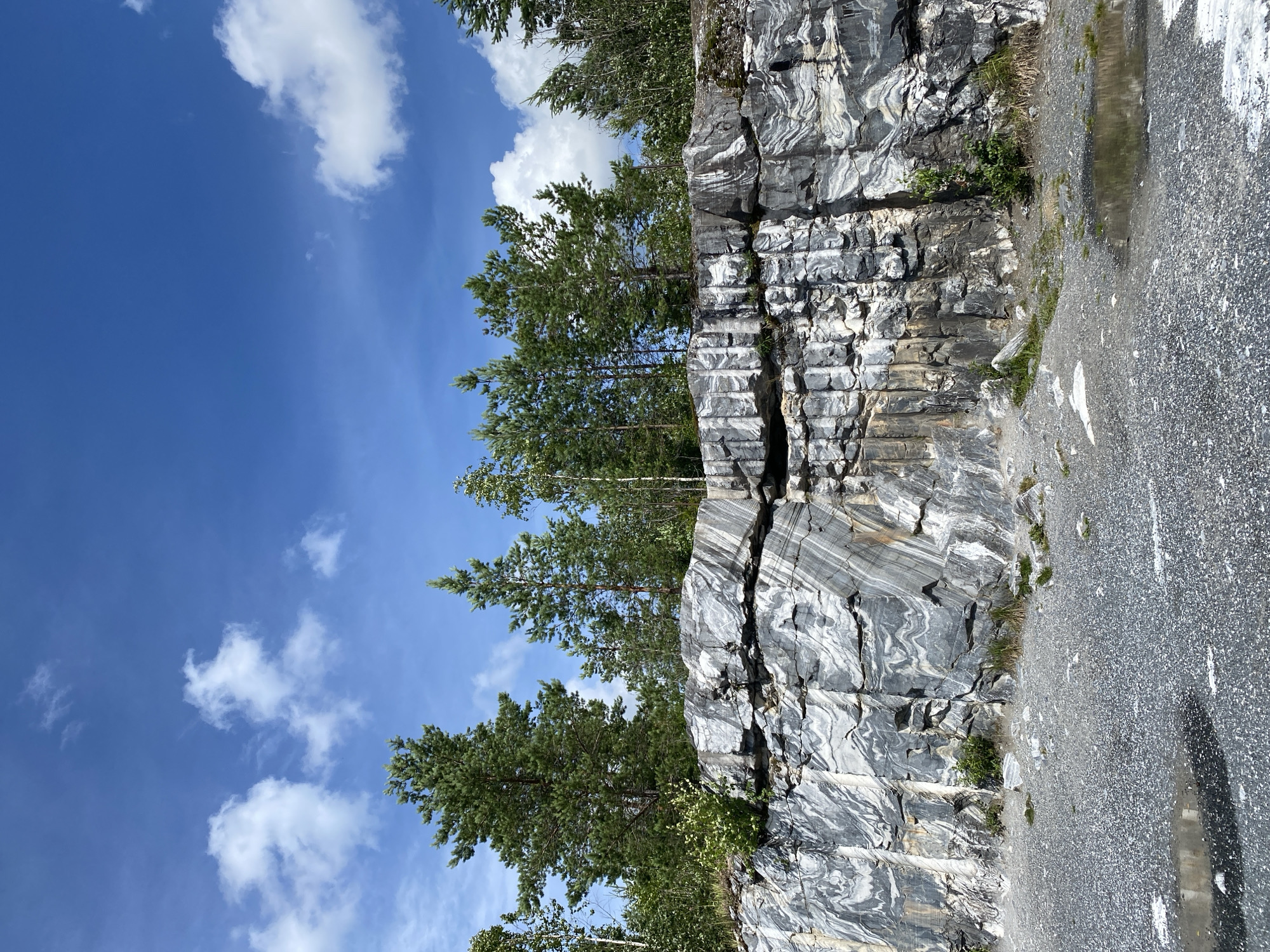 The image depicts a section of the Ruskeala Mountain Park in Ruskeala, Russia, featuring a dramatic marble cliff face under a bright blue sky with scattered white clouds. The rock formation exhibits striking gray and white banding, indicative of marble. Pine trees and other foliage grow along the top of the cliff and in small patches on its face. The ground in the foreground is covered in gravel and features several small puddles of water, reflecting the sky. The scene is a natural landscape, with no people or animals visible.