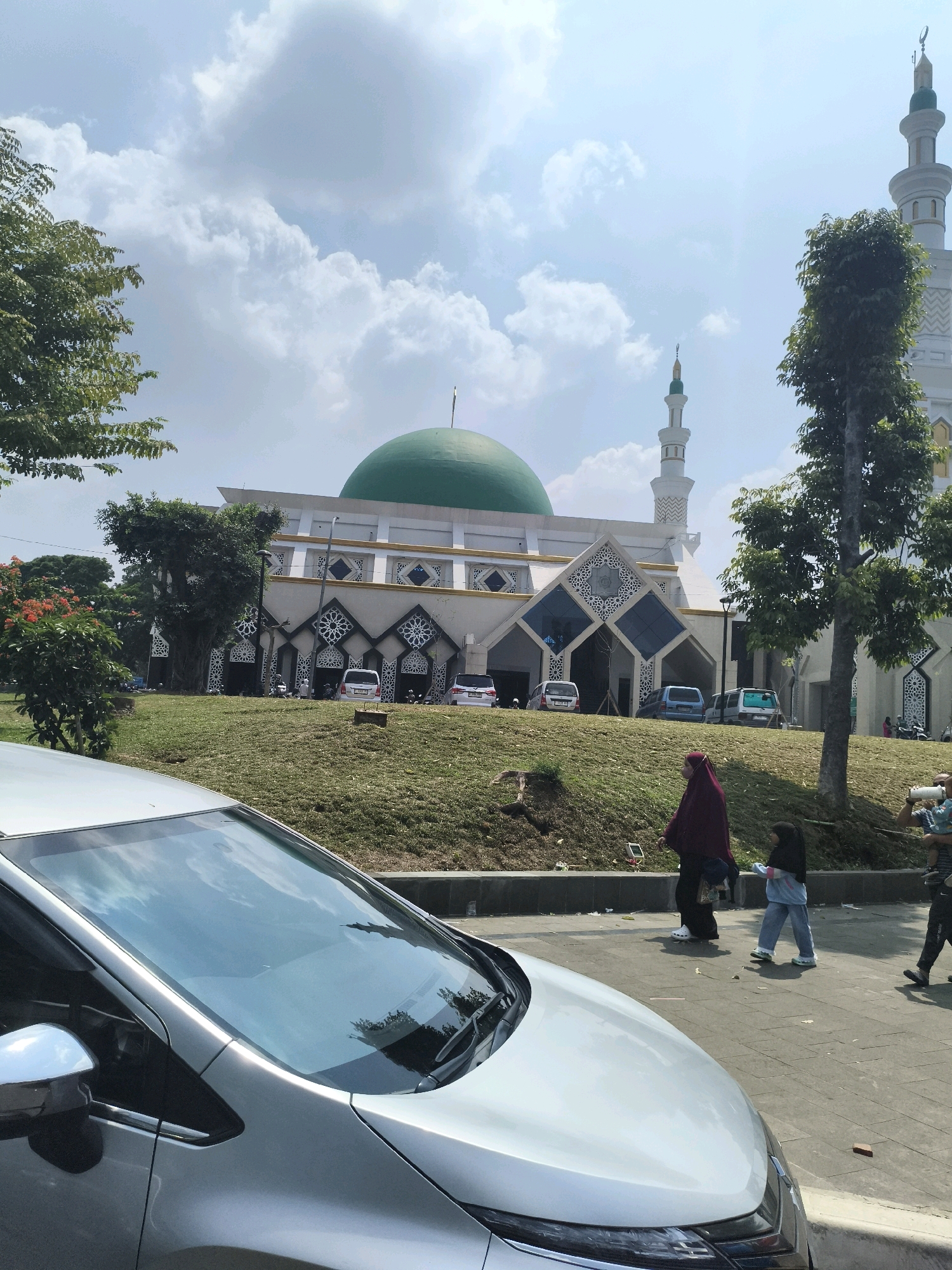 A silver car is parked in the foreground, its windshield reflecting a cloudy sky. Behind the car, a grassy slope leads up to a mosque with a prominent green dome. The mosque features white walls with intricate geometric patterns and two tall minarets. Several cars are parked in front of the mosque. A few people are visible walking on a paved walkway in front of the slope, including a woman in a maroon hijab and a young girl in blue jeans. The overall scene suggests a sunny day in Cibinong, Indonesia.