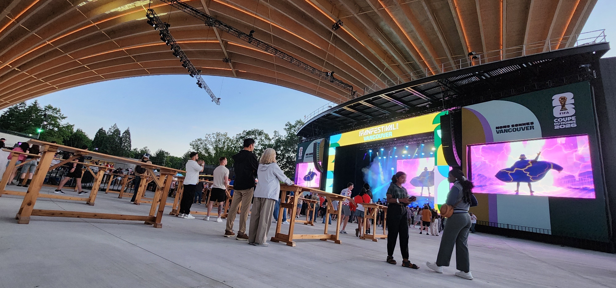 The image depicts an outdoor fan festival event, likely a FIFA World Cup celebration, as indicated by the visible text "FIFA COUPE DU MONDE 2026" and "FANFESTIVAL VANCOUVER". The scene is set under a large, curved wooden canopy, with a stage and large screens dominating the background. The stage is illuminated with colorful lights and haze, suggesting a performance or presentation is underway.
Several people are present, standing and watching the stage. Some are positioned behind wooden barriers that delineate viewing areas. The individuals are dressed casually, and their postures suggest engagement with the event. The time of day appears to be late afternoon or early evening, with the sky visible above the canopy showing a clear blue hue. The overall atmosphere seems to be one of anticipation and enjoyment.
Notable details include the modern stage setup with large LED screens displaying animated graphics. One screen shows a stylized character in a blue cape against a purple, lightning-filled background, with the text "WE ARE VANCOUVER" visible. Another screen displays "NORS SOMMES VANCOUVER", likely meaning "We are Vancouver" in French. The architectural design of the canopy, with its exposed wooden beams and integrated lighting, is also a prominent feature. The ground is paved with concrete.