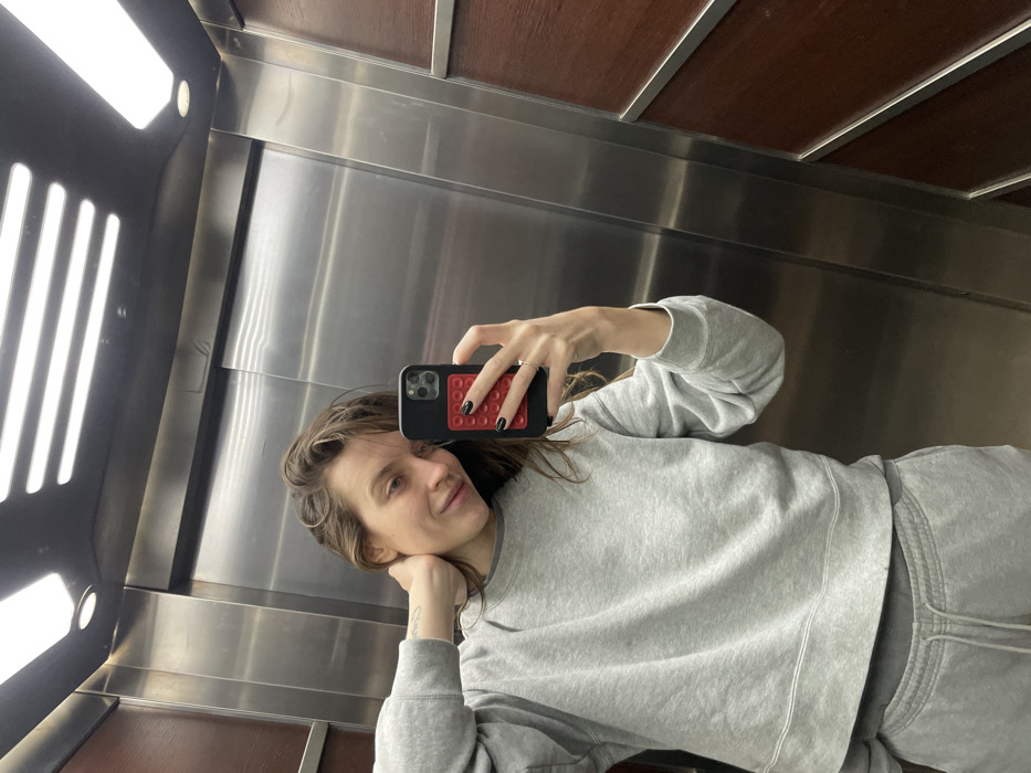 An adult female is depicted taking a mirror selfie inside an elevator located in the city of Budva, Montenegro. The elevator's interior walls are predominantly polished stainless steel, with a section of dark brown wood paneling bordered by silver trim visible on the right. Overhead, a dark ceiling houses two linear fluorescent light fixtures and one circular recessed light. The female subject, with long brown hair, is looking forward with a slight smile. She wears a grey crew-neck sweatshirt over a darker grey t-shirt, paired with matching grey sweatpants. Her right hand is positioned behind her neck, revealing a small tattoo on her inner forearm. Her left hand holds a black smartphone, which features a red, textured attachment on its back. The subject's fingernails are painted black, and a ring is worn on her left ring finger.