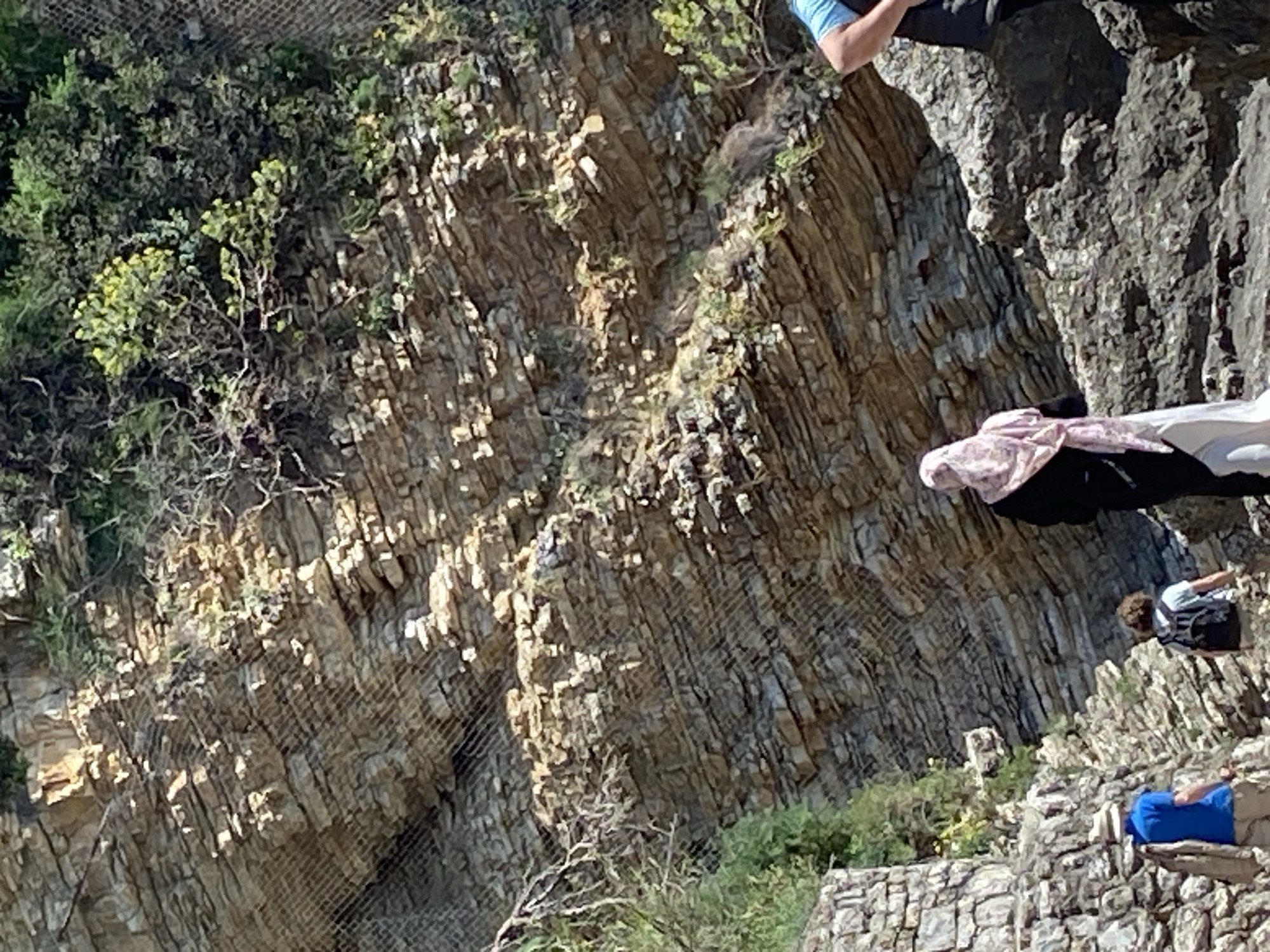 The image shows a rock face with a wire mesh covering a portion of it, likely for erosion control. Some vegetation clings to the rock, including greenery and small trees. Several people are positioned on different levels of the rocks: one is standing on a higher level with part of their legs visible, another is standing on a lower rock formation, and a third is seated on the rocks at the bottom of the image. The location is Sveti Stefan, Montenegro.