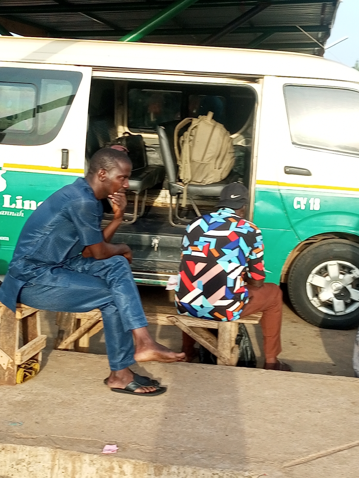 Two men are sitting on wooden benches outside a white and green bus, seemingly in a bus station. The man on the left, wearing a blue striped outfit, is facing left and resting his chin on his hand. The man on the right, wearing a black cap and a colorful geometric-patterned shirt, is facing away from the camera, looking towards the bus. A beige backpack is visible inside the bus, near the open doorway. The bus has "CY 18" written on its side. The ground appears to be concrete. The scene is lit by natural daylight. This scene is characteristic of a bus terminal in Gombe, Nigeria.