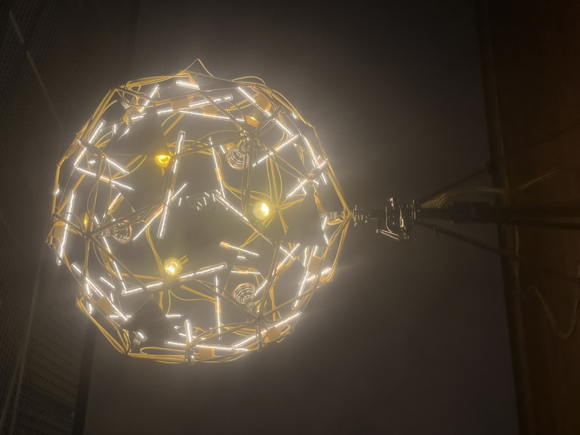 A low-angle, close-up shot presents a spherical light fixture against a predominantly dark backdrop. The light fixture is constructed from a yellow framework intertwined with numerous white, linear lights, creating a complex and intricate design. Four visible traditional incandescent light bulbs with yellow light add to the overall luminosity of the structure. To the right, a mechanical arm supports the sphere, extending horizontally. The location is not evident in the image.