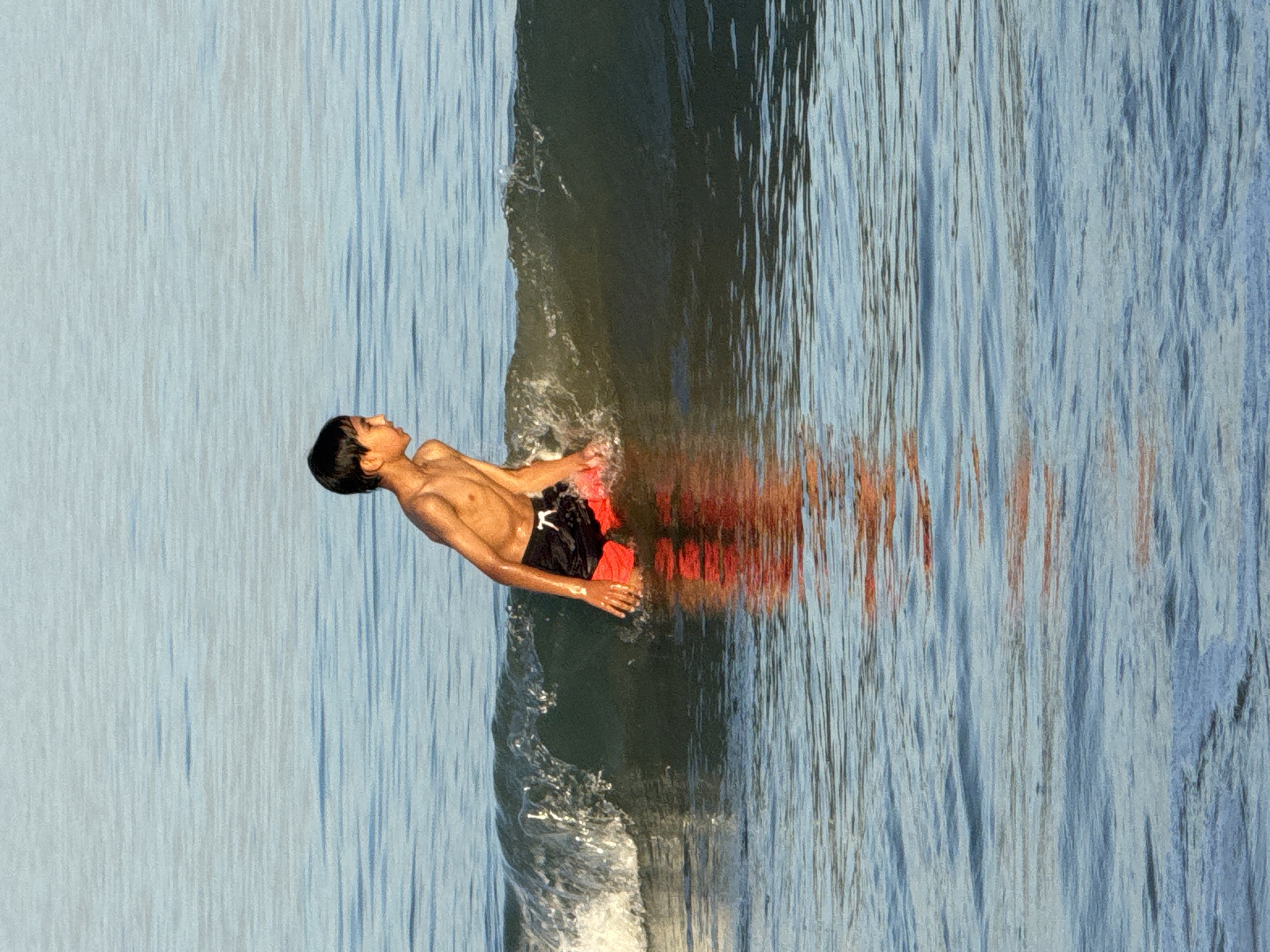 A tanned boy in black and orange shorts floats horizontally on the surface of water. His reflection, distorted by the water, mirrors his form beneath him. A gentle wave breaks around his midsection. The water is surrounded by a blue texture, resembling the sky. This appears to be a photograph taken at Jimbaran Beach, Indonesia.
