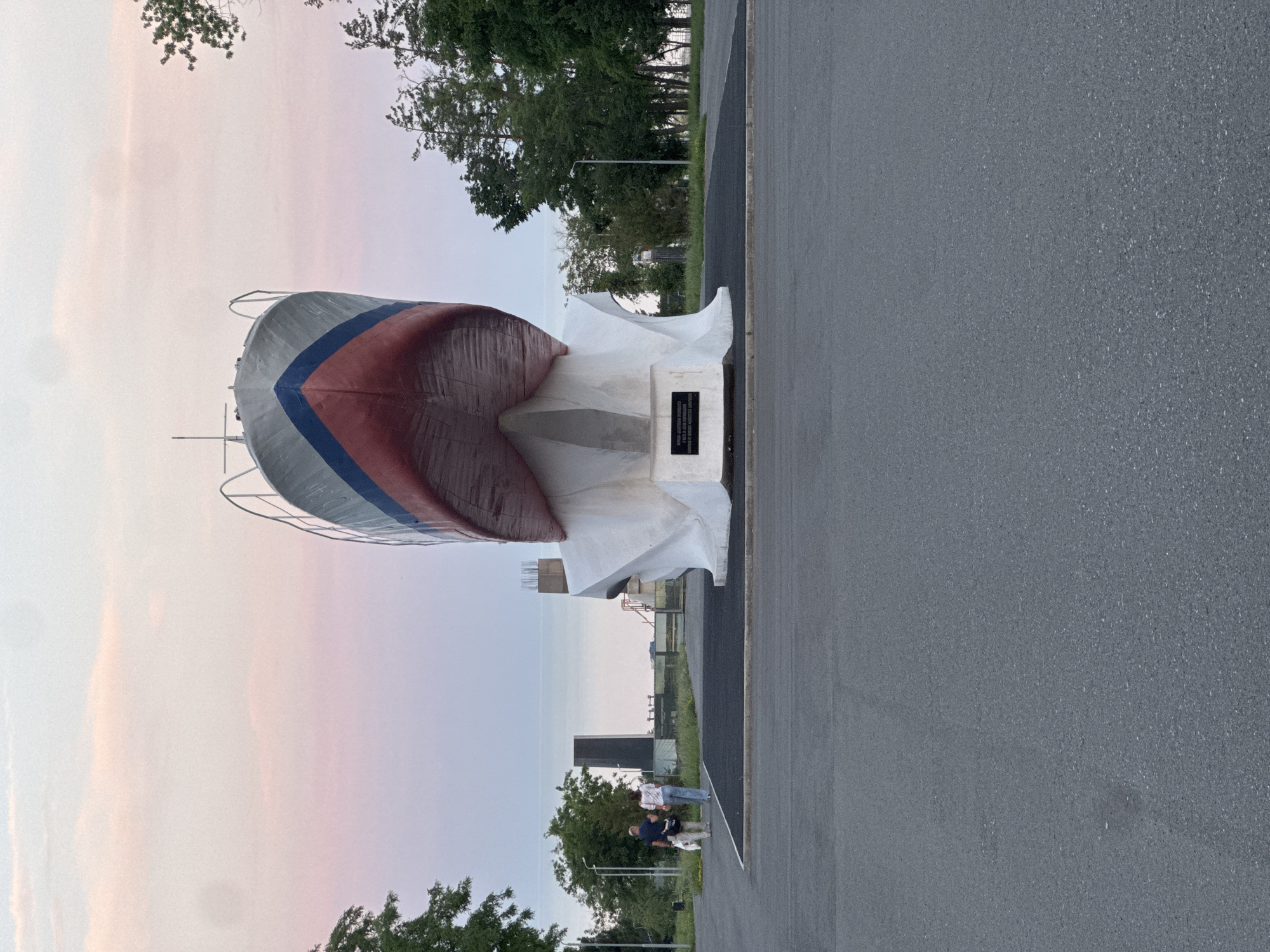 This is a full shot of the "Sail of Victory" monument in Mariupol, Ukraine, a memorial dedicated to the city's liberation from Nazi occupation. The monument depicts the bow of a ship, painted red, white, and blue, mounted on a white pedestal. The ship is angled upwards, with the hull facing the camera. The monument is situated on a paved area with a dark grey asphalt road in the foreground. Two people, a man and a woman, walk away from the camera towards the monument. They are dressed casually, with the woman carrying a bag and the man holding a white plastic bag. A small white dog walks beside them. In the background, there are trees, streetlights, and a dark, rectangular obelisk. The sky above is a soft gradient of pink and blue, suggesting either dawn or dusk. The monument is located at the intersection of Prospekt Metallurgov and Nakhimova Avenue in Mariupol, Ukraine.