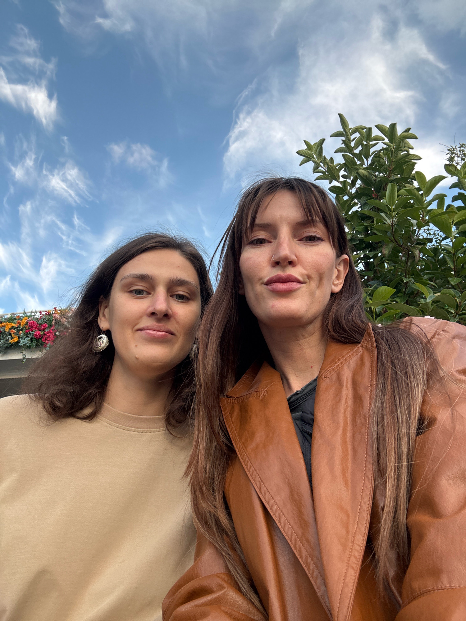 Two women are standing outdoors, posing for a selfie. The woman on the left has long, dark, wavy hair and is wearing a tan crewneck shirt and large, ornate earrings. The woman on the right has long, dark, straight hair with bangs and is wearing a brown leather jacket over a black garment. They are both looking at the camera. Behind them, the sky is visible with wispy white clouds, and a green tree with dense foliage. There is also a railing with colorful flowers in the background on the left. The scene appears to be during the daytime.
