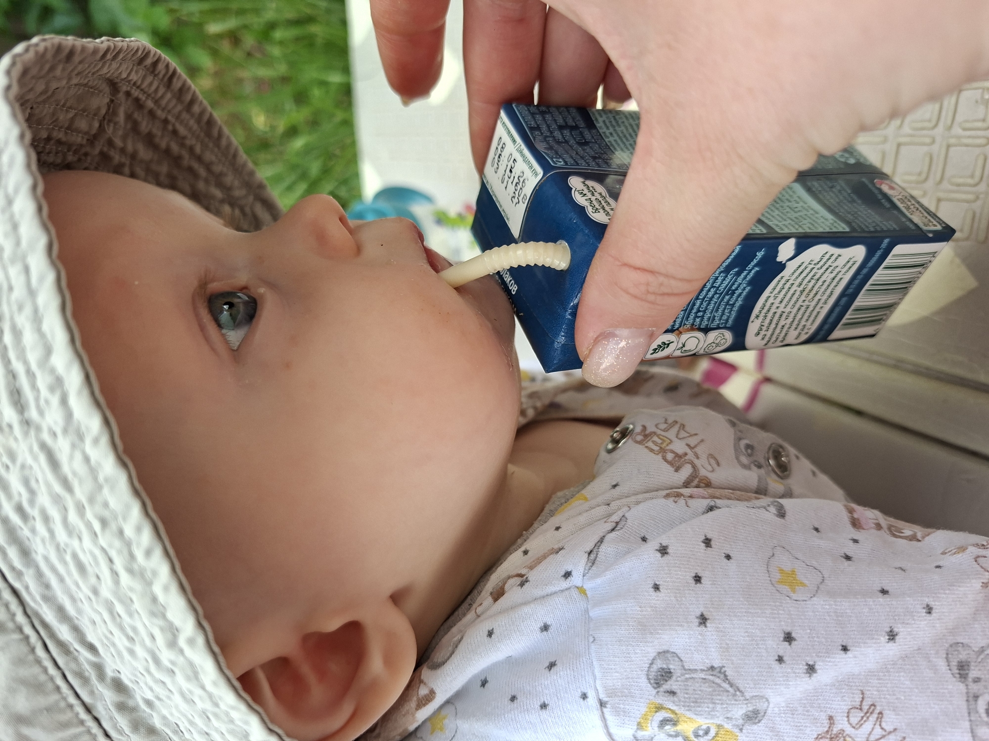 A close-up, profile shot shows a baby wearing a light-colored bucket hat and a white onesie with gray teddy bears and yellow stars printed on it. The baby is looking to the right, with one eye visible. A person's hand, with a light pink manicure, is holding a small blue juice box with a white straw. The straw is placed in the baby's mouth. The background is blurred green grass.