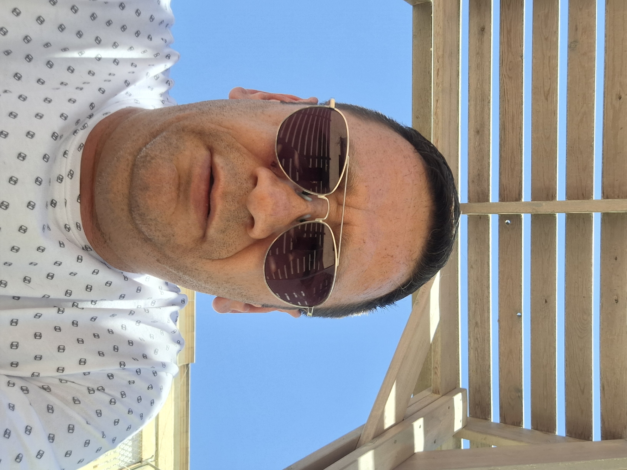 The image is a close-up, low-angle selfie of a man in Yerocham, Israel. He is wearing sunglasses and a white t-shirt with a small, repeated pattern of what appear to be cubes. The man is outdoors, under a wooden pergola, with a clear blue sky visible through the slats of the structure. The pergola's shadow is cast across the man's face and shirt. The background shows the bright sky and a hint of a light-colored building structure, possibly a fence or wall, with a yellow awning.