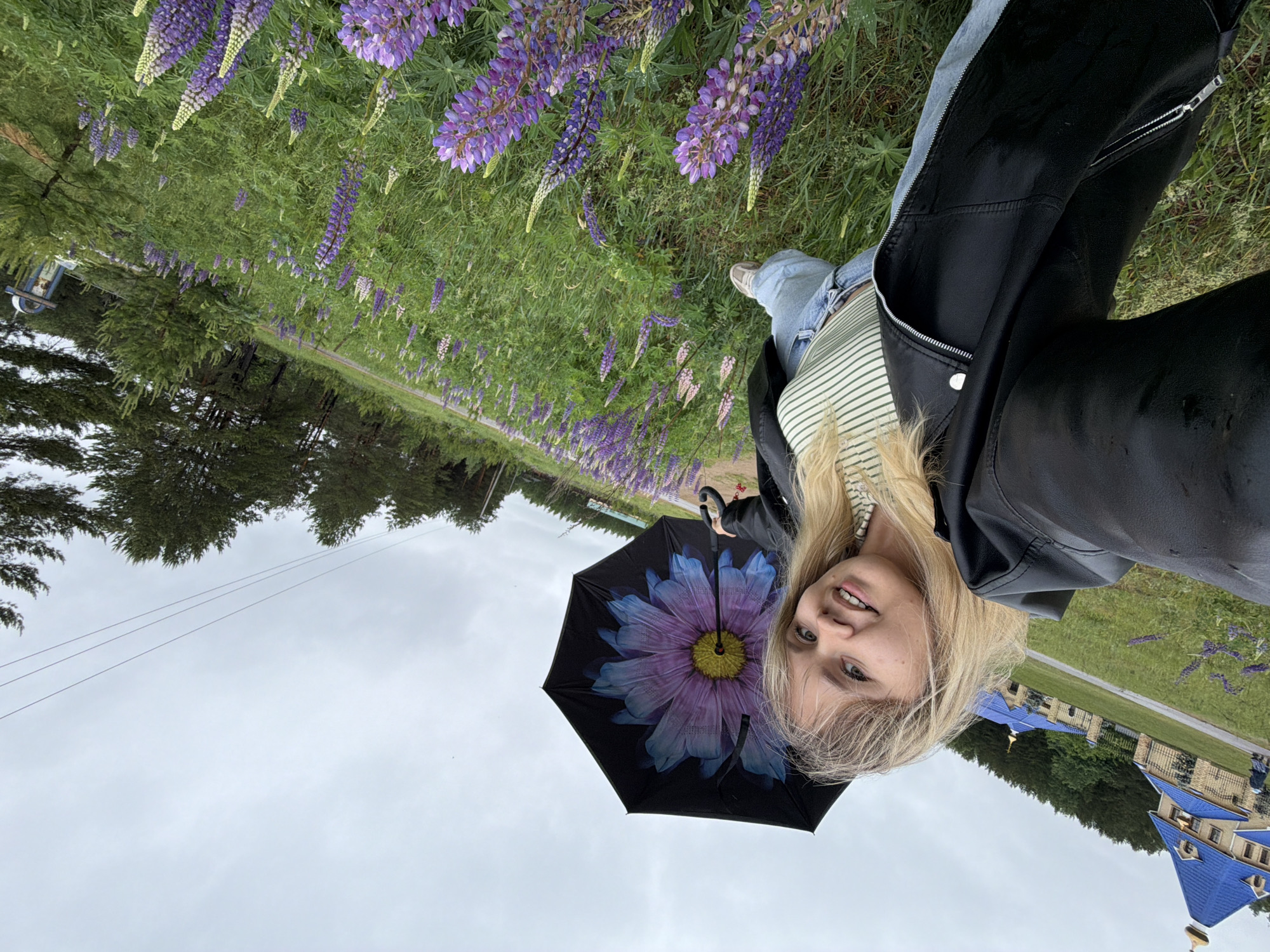 A photograph shows a light-skinned woman holding a black umbrella with a large, colorful flower design in front of her face. She is wearing a black leather jacket, a striped shirt, and jeans. The background shows a field of purple lupine flowers, trees, a cloudy sky, and a building with a blue roof and gold domes. The building is the Holy Spring and Church of St. Sergius of Radonezh in Chaikovsky, Russia.