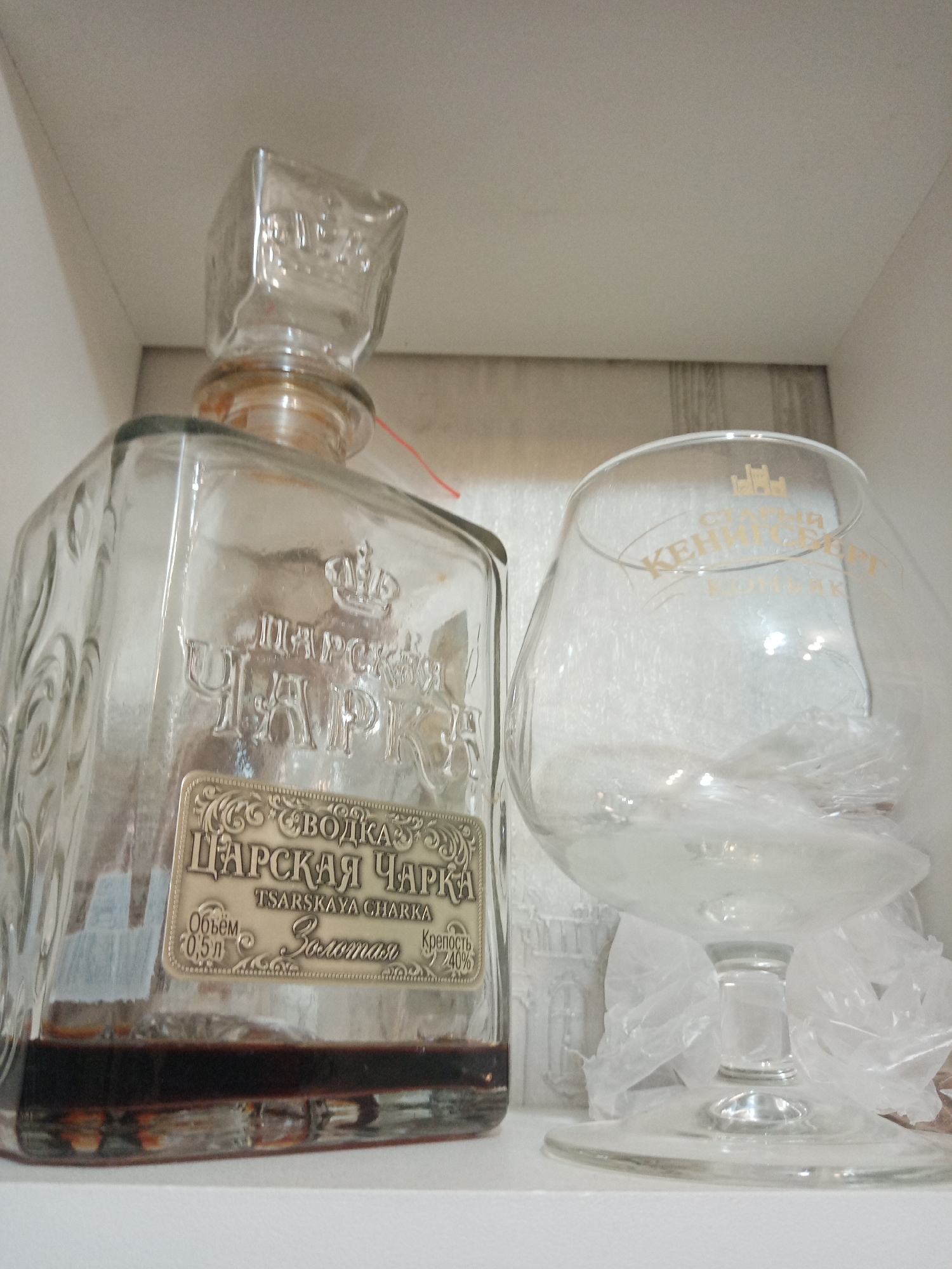 A glass bottle labeled "Tsarskaya Charka" (Царская Чарка) with its matching square stopper and a snifter glass with a logo are on a white surface. The bottle is approximately 1/5 full with a dark liquid and is marked with a label indicating it is a vodka with 40% alcohol and 0.5L volume. The snifter glass has packaging inside. The scene is most likely in Elektrostal, Russia, judging by the Russian text on the bottle.