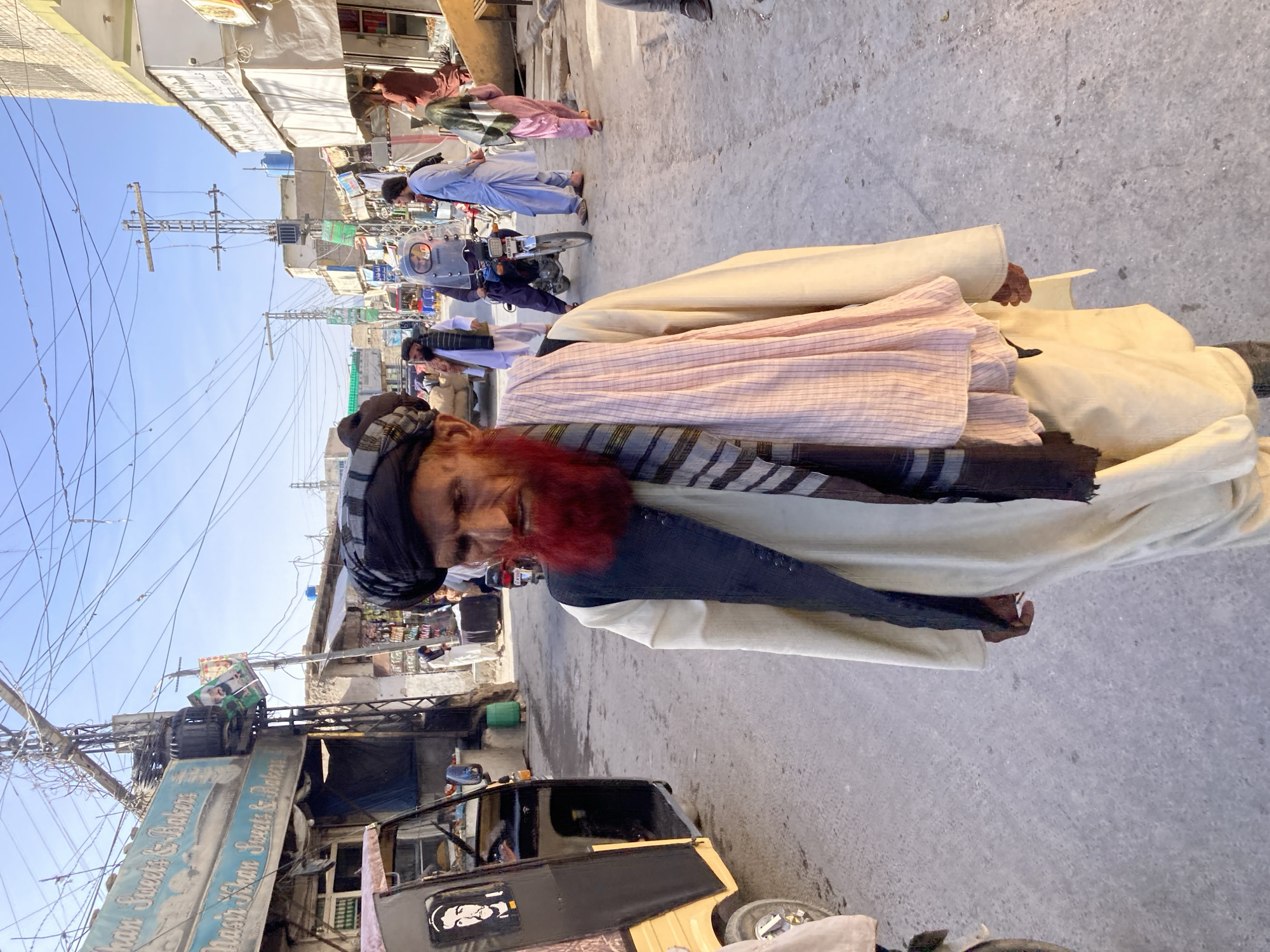 A medium, outdoor shot depicts a man with a red-dyed beard and head covering standing in a street in Quetta City Tehsil, Pakistan. He is wearing a tan and white robe with a striped scarf. The setting appears to be a busy street with buildings, power lines, and other people in the background. A yellow and black auto rickshaw is parked in the foreground.