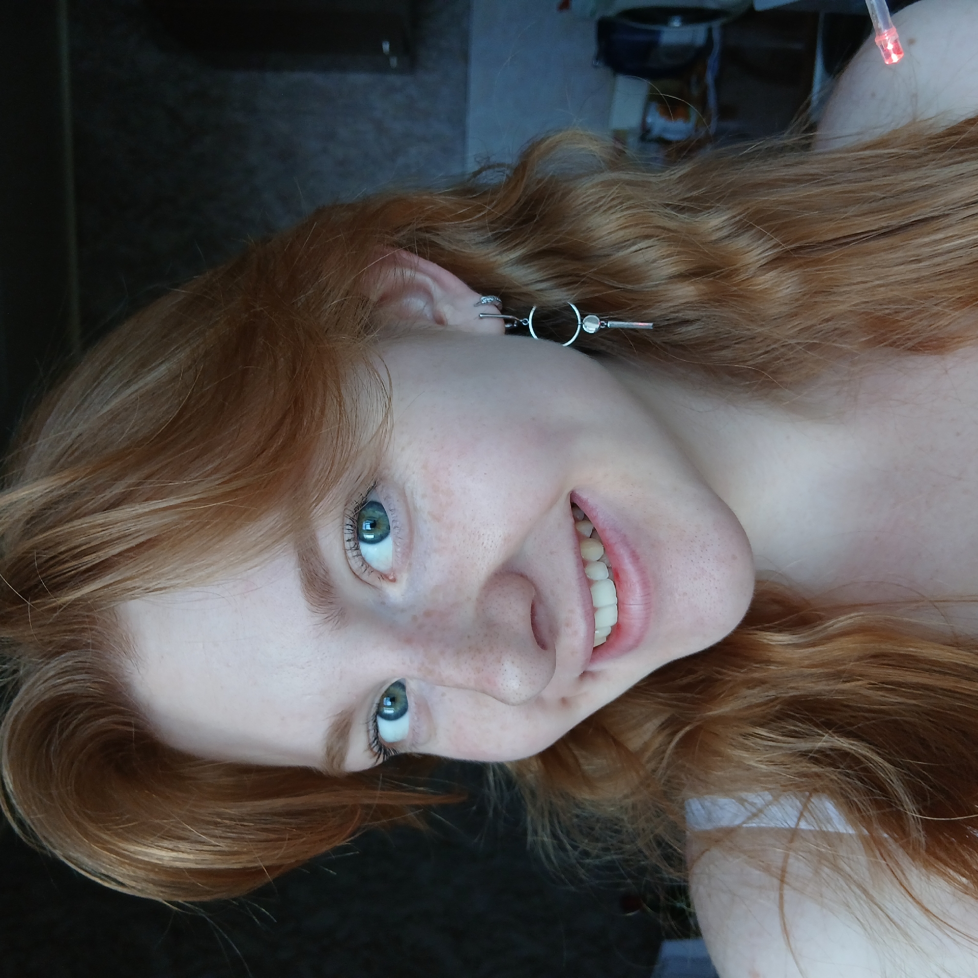 A close-up portrait, originally rotated 90 degrees clockwise, shows a young person with fair skin and numerous freckles on their nose and cheeks. They have long, wavy red or auburn hair and striking blue-green eyes. The person is smiling broadly, revealing their teeth, and looking directly at the camera, conveying a cheerful and happy emotion.
They are wearing silver-toned earrings. One ear features a distinctive earring composed of two horizontal bars connected by two circular elements, while the other ear has a simple hoop earring.
The background suggests an indoor setting, with a dark, indistinct area to the left and a lighter, somewhat cluttered space in the upper right. A small, bright red glowing object, possibly a light from an electronic device or a pointer, is visible in the upper right corner, near the person's shoulder. No specific location cues, time of day, or visible text are present.