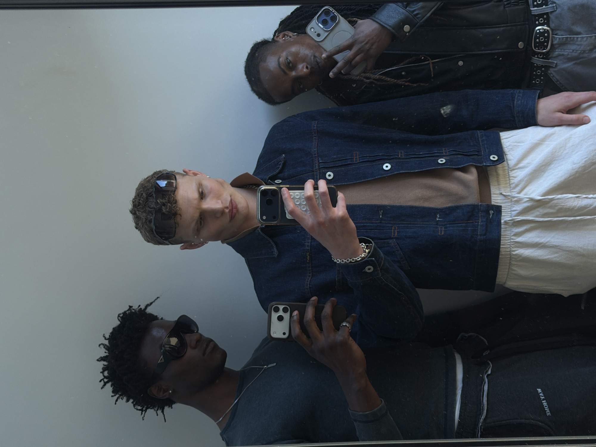 The image captures a mirror selfie featuring three young men, each holding an iPhone. The scene is presented as if viewed from above, with the individuals reflected and rotated clockwise.
In the upper right of the reflection, a person with dark skin and dark braided hair looks directly at the camera. They wear a dark jacket, possibly leather, and multiple ear piercings. They hold a light-colored iPhone with a dark case.
Centered in the reflection is a person with light skin and short, curly light brown hair, wearing sunglasses pushed up onto their head. This individual is dressed in a dark blue denim jacket over a light brown top and white, relaxed-fit pants. A silver chain bracelet adorns their wrist. They hold a dark-colored iPhone, appearing to look at its screen.
In the lower left of the reflection, another person with dark skin and short, curly dark hair is visible. They wear dark sunglasses and a dark long-sleeved top, accessorized with a silver chain. This person also holds a dark iPhone, seeming to look upwards or towards their phone.
All three individuals are reflected against a plain, light gray background, indicating an indoor setting. The mirror surface shows several small dust particles. The iPhones are modern models, identifiable by their multiple rear camera lenses. On the dark top worn by the person in the lower left, the text "MYA HOME" is visible in reverse due to the mirror's reflection. The overall atmosphere is casual, and the activity is posing for a photo.