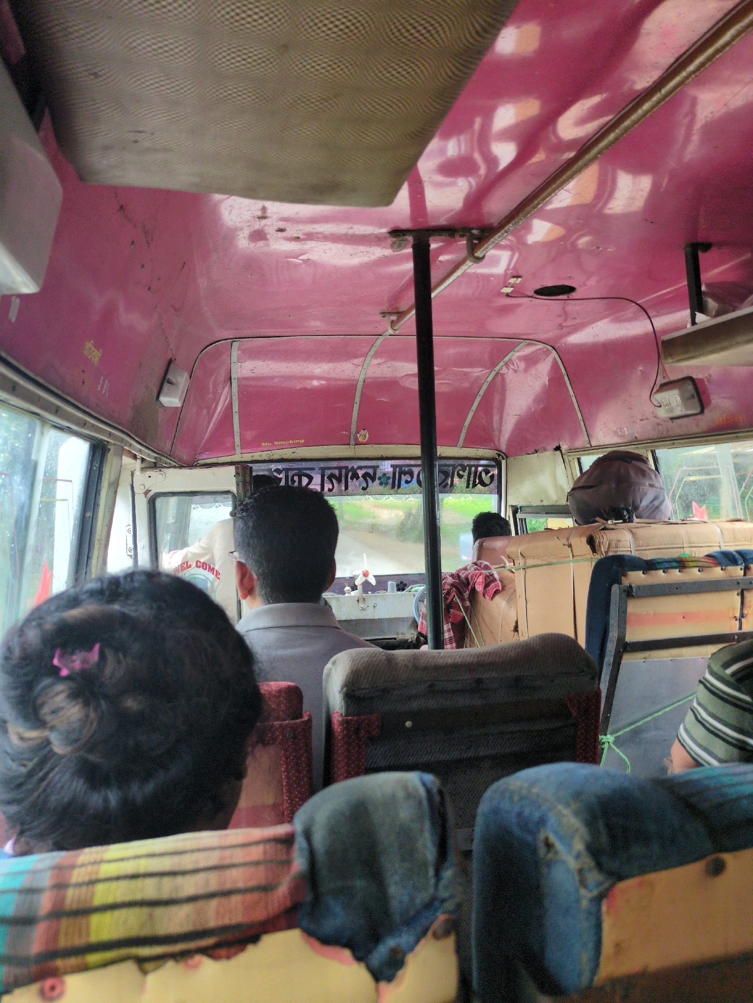 The image is an interior view of a bus, likely in Teliamura, India, looking towards the front. The ceiling of the bus is painted a bright pink and shows signs of wear. A metal pole runs vertically in the center of the aisle. The windshield at the front reveals a rural, green landscape. Inside the bus, several passengers are visible. In the foreground, the back of a woman's head with dark hair and a pink hair accessory is prominent. Further forward, a man in a grey shirt is seated, facing away from the camera, with glasses visible. Behind him, another passenger, possibly a man, is seated, with a dark backpack or bag on his lap and shoulders. Cardboard boxes are stacked behind some seats. The seats themselves appear worn, with some covered in fabric and others with exposed padding. A green rope is tied around some of the seat backs. Another person in a green and white striped shirt is partially visible on the right side. The overall impression is of a crowded and functional bus environment.