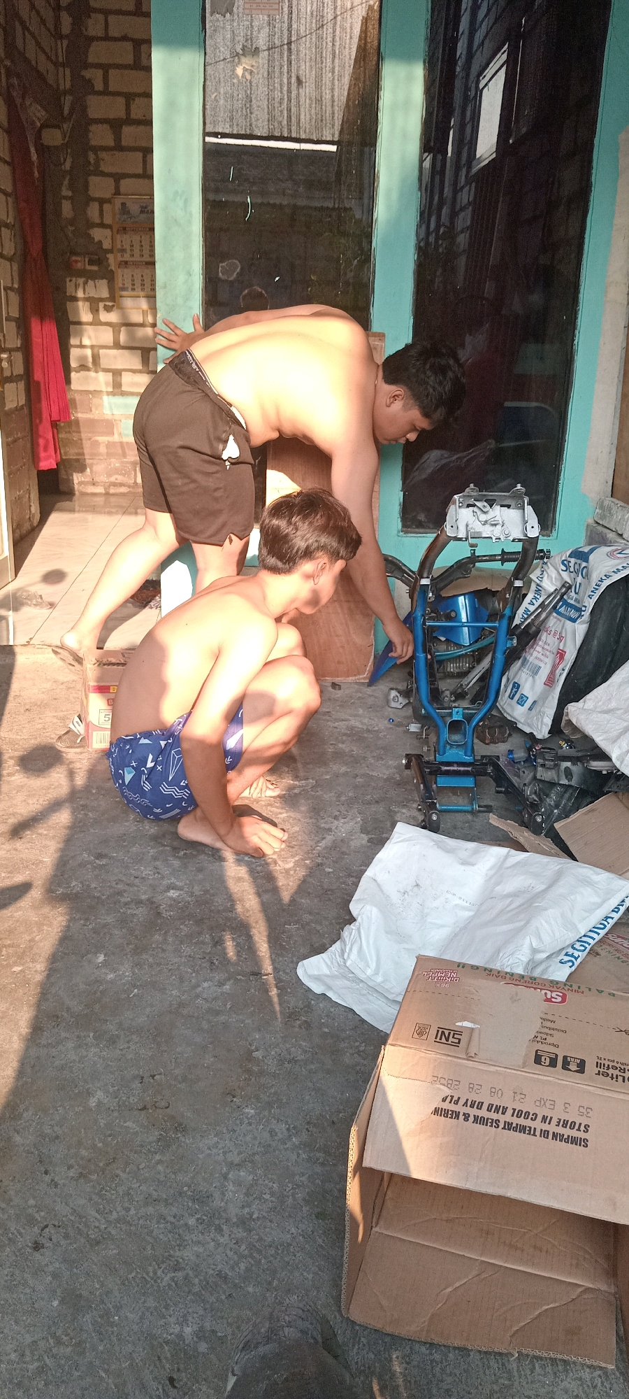 Two shirtless young men are engaged in an activity outdoors in what appears to be a backyard or alley in Babat, Indonesia. The man on the left is crouched down, wearing blue patterned shorts, and is looking at the ground. The man on the right is standing and leaning forward, with his hands near a disassembled blue metal frame, possibly part of a motorcycle or bicycle. He is wearing black shorts. The environment is cluttered with various objects, including cardboard boxes, plastic bags, and building materials, suggesting a work or storage area. The lighting indicates it is daytime, with strong shadows cast on the concrete ground. A brick wall and a building with a glass-paneled door are visible in the background.