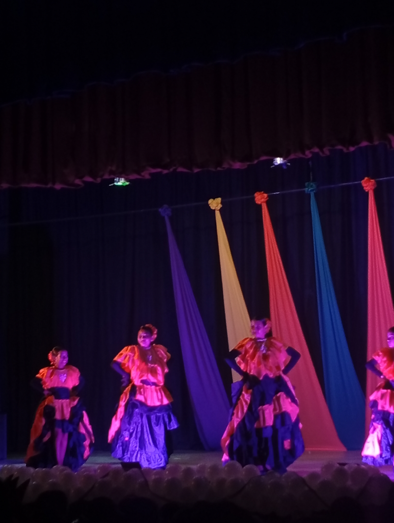 The image captures a dance performance in what appears to be a theater or auditorium, potentially in Juan Griego, Venezuela. The stage is set with a dark curtain backdrop adorned with colorful hanging sashes of yellow, orange, blue, and purple. Four women are dressed in vibrant costumes of orange and black, which feature full skirts and ruffled detailing. The women are positioned across the stage, appearing to be in the midst of a choreographed dance, as indicated by their posed stances and focused expressions. The stage floor seems to be covered in what looks like white balloons. The setting is illuminated by stage lighting, casting a theatrical atmosphere. The image's composition emphasizes the performance aspect of the event.