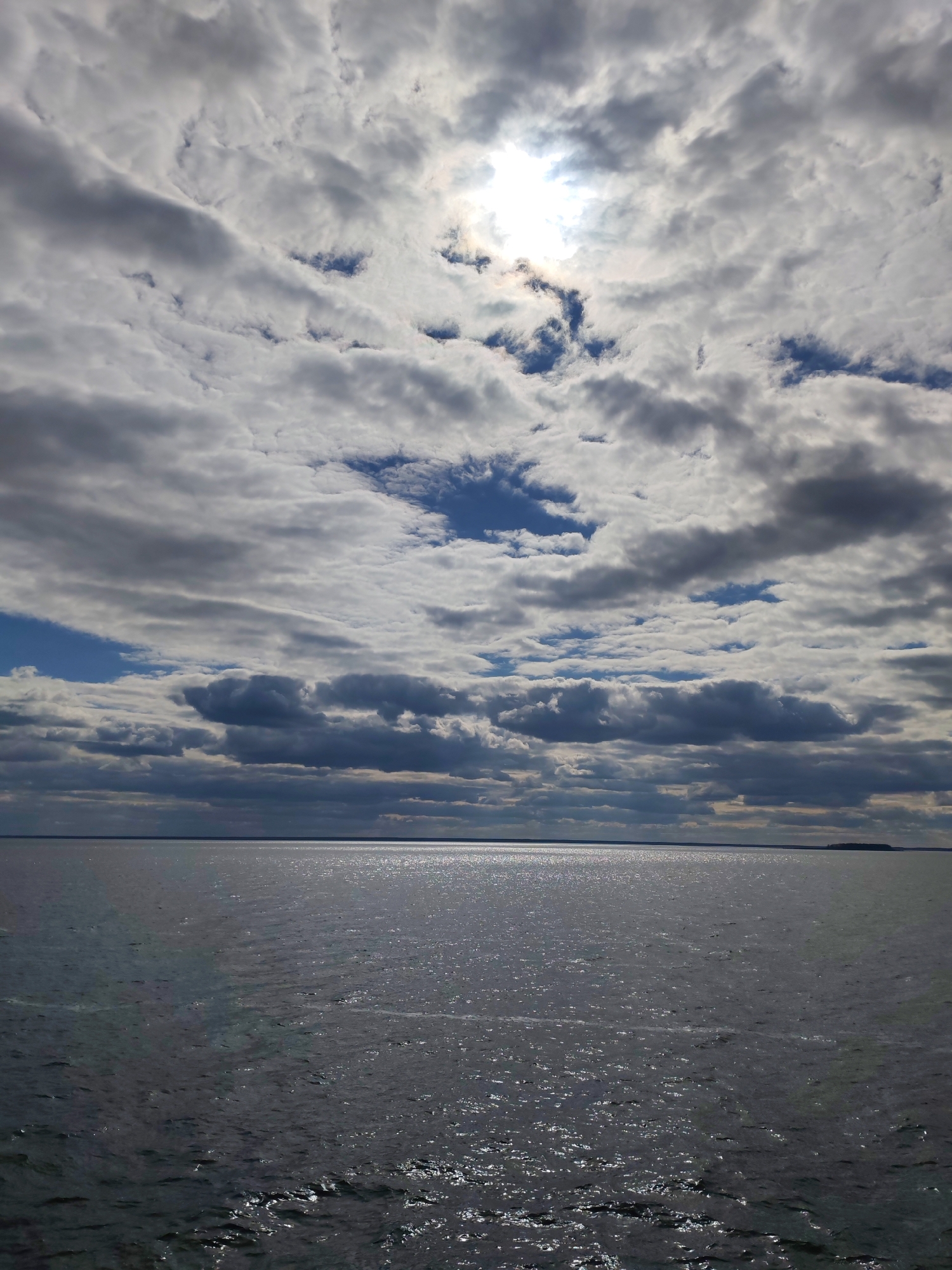 The image captures an expansive outdoor scene featuring a vast body of water under a dynamic, cloudy sky. The upper two-thirds of the frame are dominated by a dense and varied cloud cover, consisting of a mix of stratocumulus and cumulus clouds. Patches of bright white and light gray clouds are interspersed with darker, denser cloud formations. The sun shines brightly through a significant break in the clouds in the upper-center, creating a strong, radiant glow. Several smaller patches of clear blue sky are visible between the clouds, especially towards the left and central regions of the sky.
The lower third of the image shows a wide expanse of dark, rippling water, likely a sea, large lake, or river. The surface of the water reflects the light from the sky, with a shimmering, glinting path of sunlight stretching across it directly beneath the sun. This bright reflection creates a strong contrast with the darker surrounding water. The water extends to a distant, flat horizon line, with a faint, dark line on the far right suggesting a remote landmass or island.
No people or distinct man-made objects are visible. The time of day appears to be late morning or afternoon, indicated by the sun's position and the strong reflections on the water. The weather is partially cloudy with periods of bright sunlight. The scene conveys a sense of vastness and natural beauty, with a dramatic interplay of light and shadow across the sky and water. There is no visible text in the image.