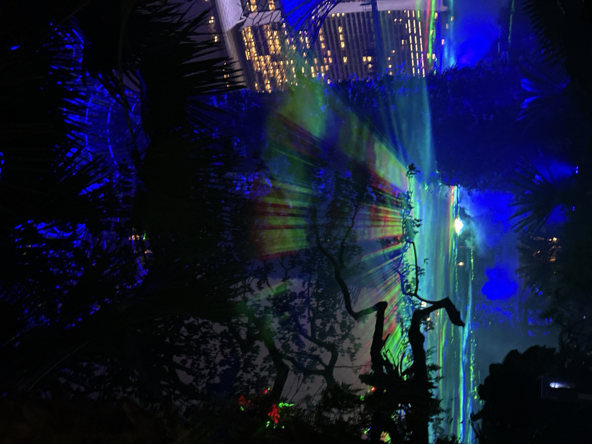 The image shows a nighttime scene at the Supertrees Grove at Gardens by the Bay in Singapore. Silhouetted trees and palm fronds are in the foreground, with vibrant, multicolored lights projected onto them and the surrounding area. The lights create a dramatic, ethereal effect, with beams of green, blue, red, and yellow fanning out across the scene. In the background, the illuminated facade of a modern skyscraper, likely part of the Marina Bay Sands complex, is visible, its windows glowing with warm light. The overall impression is one of a technologically enhanced, futuristic urban landscape, juxtaposed with natural elements.