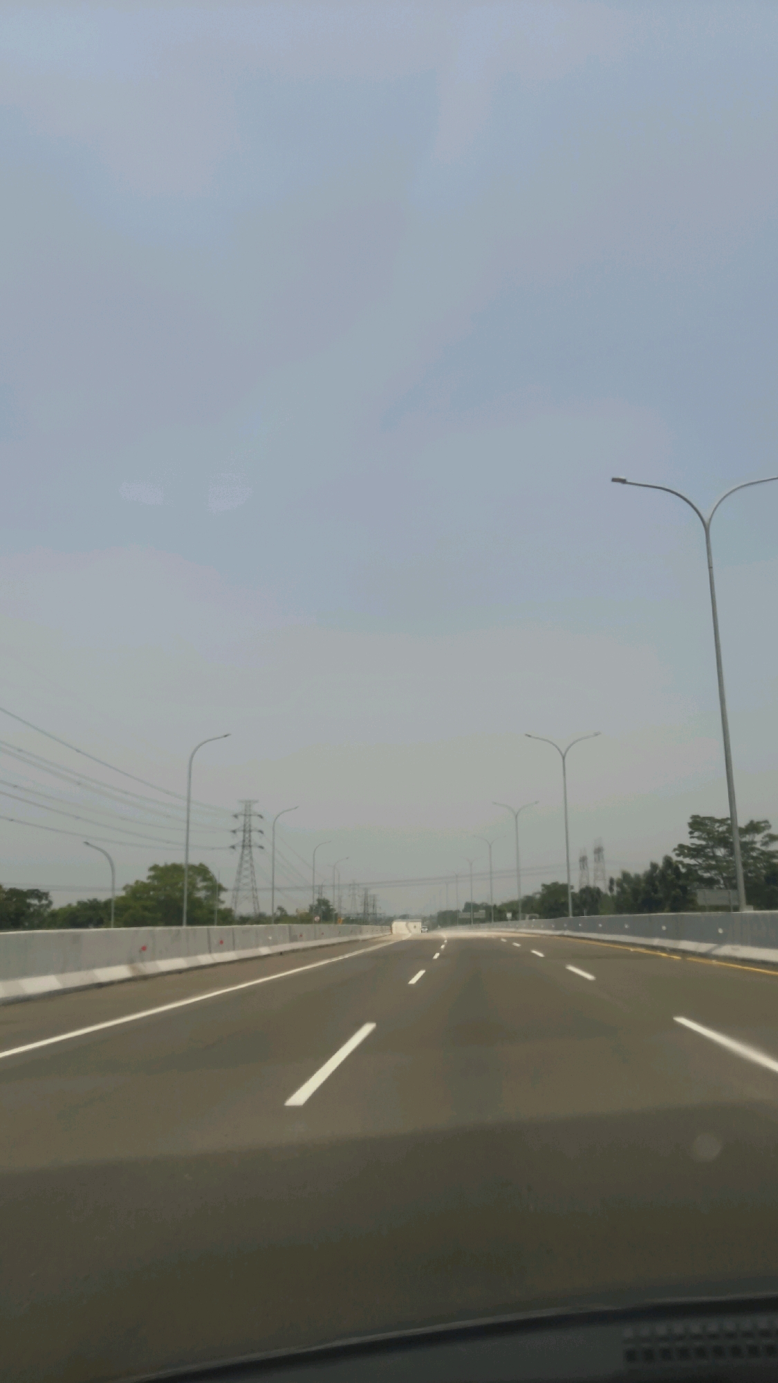 The image captures a forward-facing view from a vehicle traveling on a multi-lane highway in Situ Gadung, Indonesia. The highway is paved with asphalt and marked with white lane dividers. Concrete barriers line the sides of the road. On the left, there are power lines and pylons, as well as trees and other vegetation. Streetlights are visible on both sides of the highway, spaced at regular intervals. The sky is overcast, with a hazy, light blue-grey appearance. The overall scene depicts an infrastructure development in a semi-urban or rural area, characterized by a modern highway and associated utilities. No people or other vehicles are clearly visible in the immediate foreground or midground.