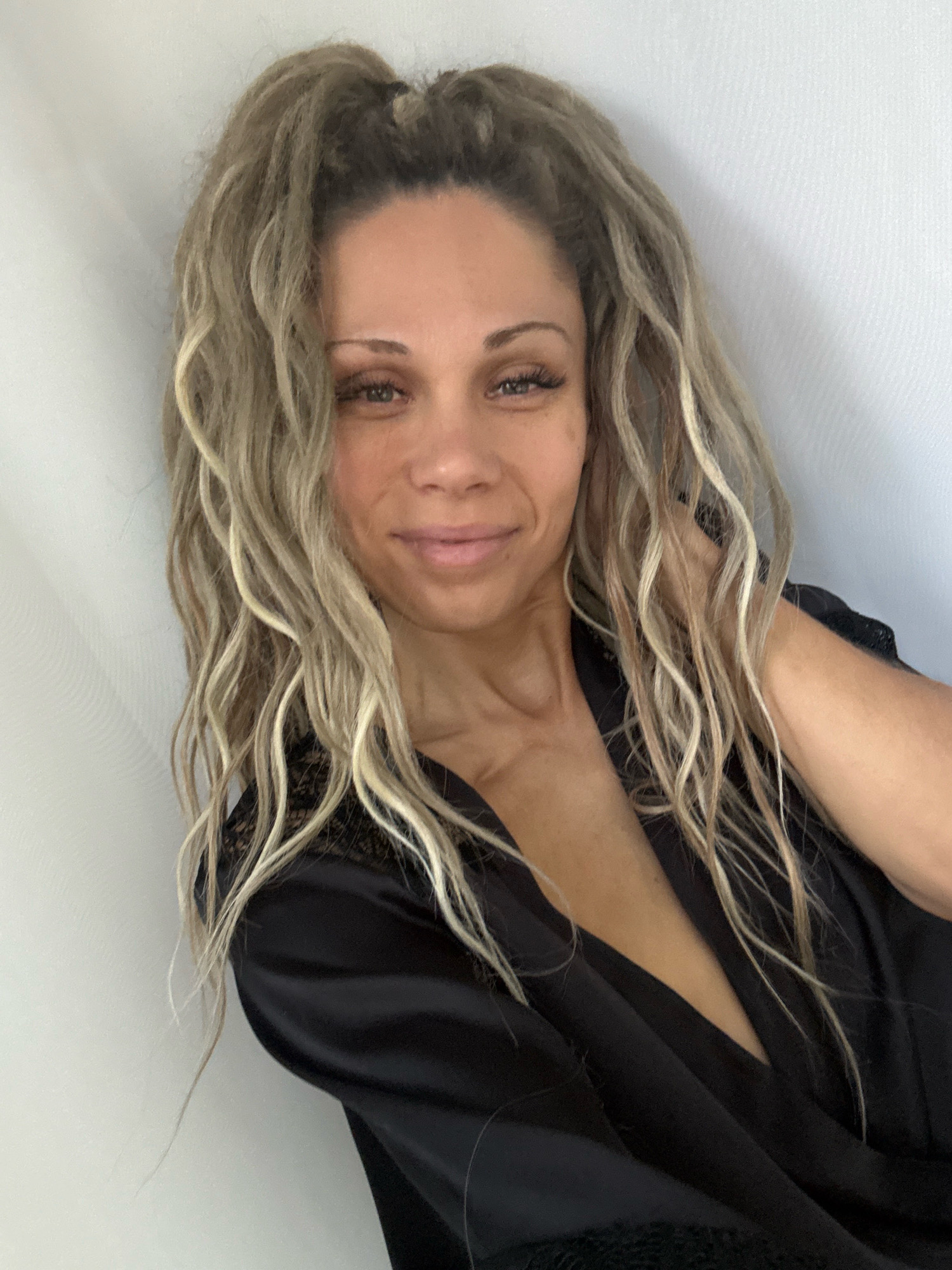 A woman with long, blonde, curly hair is seen in a close-up shot. She is wearing a black satin robe with lace detailing. The background is a plain white wall. The woman is looking towards the camera with a slight smile. Her right hand is resting on her neck. The image does not provide any information about the city of Rusanovo, Russia, or any specific location.