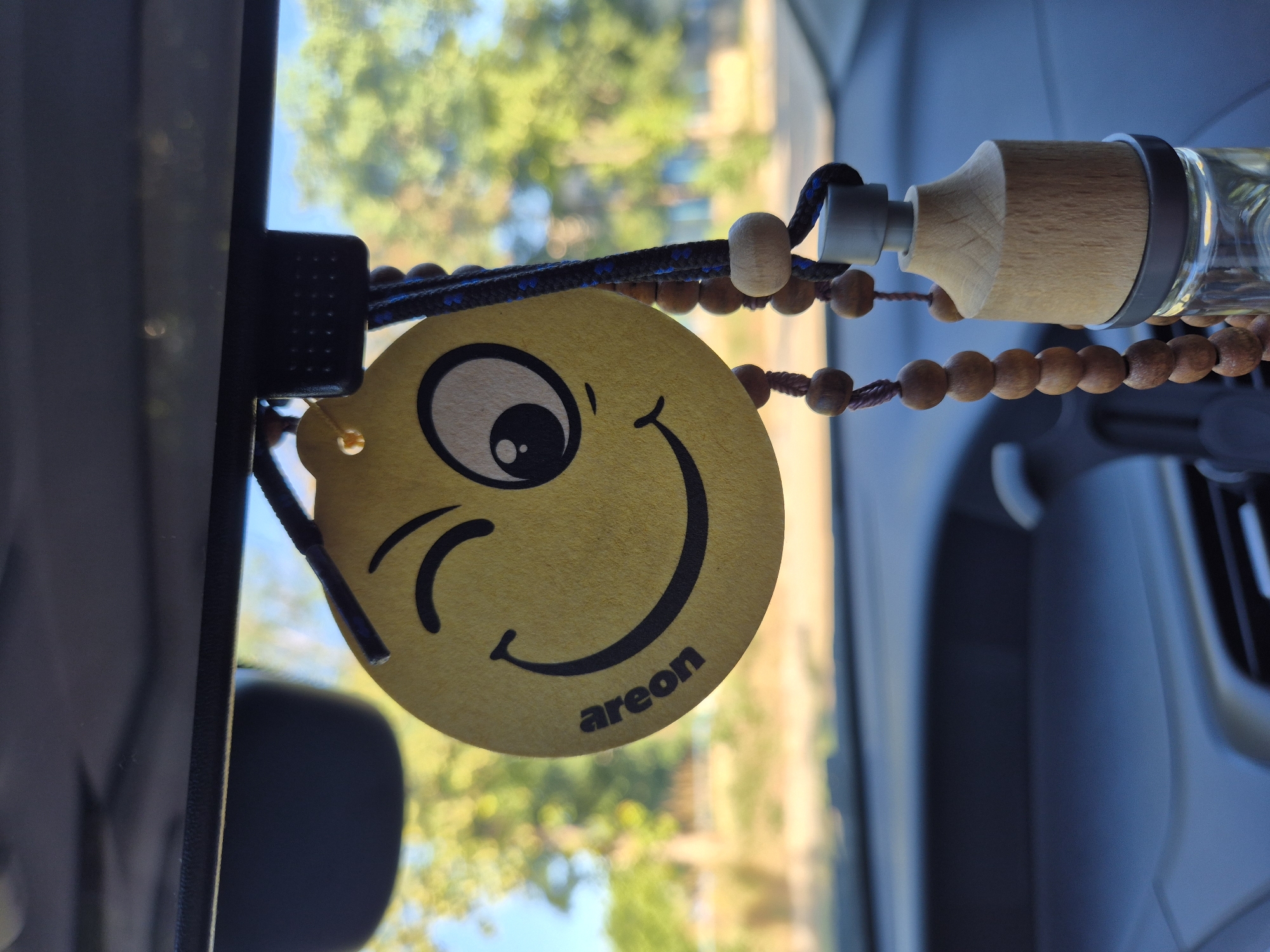 A yellow, circular car air freshener with a winking smiley face design hangs from the rearview mirror of a car. The air freshener has "areon" printed on its surface. Below the air freshener, a string of brown wooden beads hangs, attached to a small glass bottle with a light-colored wooden cap. The background of the image is blurred, showing green trees and a blue sky, suggesting an outdoor environment outside the car. The interior of the car, including the dashboard, is visible in the foreground and blurred background.