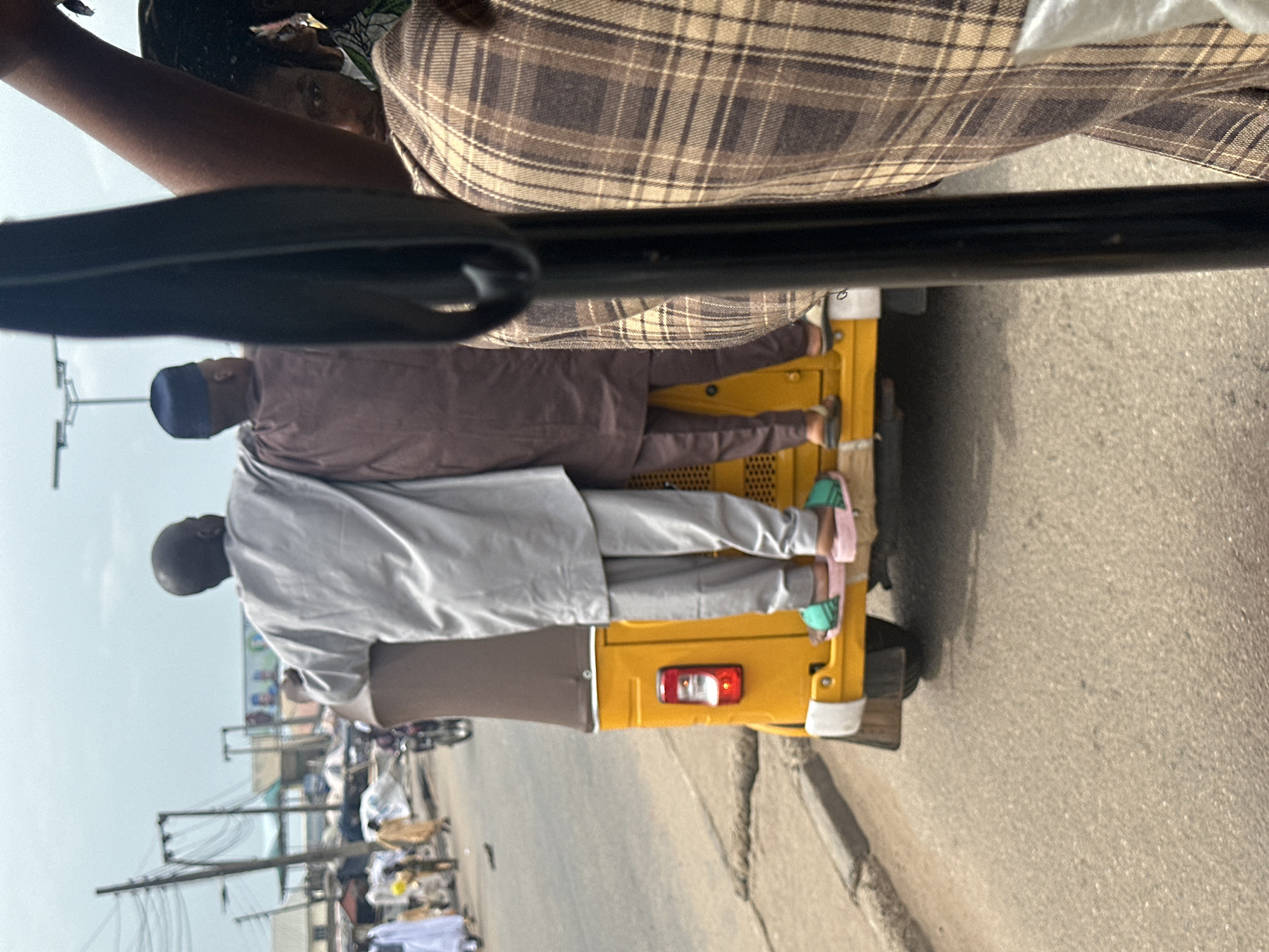 A photograph shows the back of a yellow tricycle (keke) moving on a street in Lokoja, Nigeria. Two men are seated in the back, dressed in traditional attire: one in light gray and the other in brown. The street is paved and shows signs of wear and tear. In the background, there are buildings and electrical poles. The photo is taken from the perspective of someone riding in front of the tricycle, with a black handlebar partially visible.
