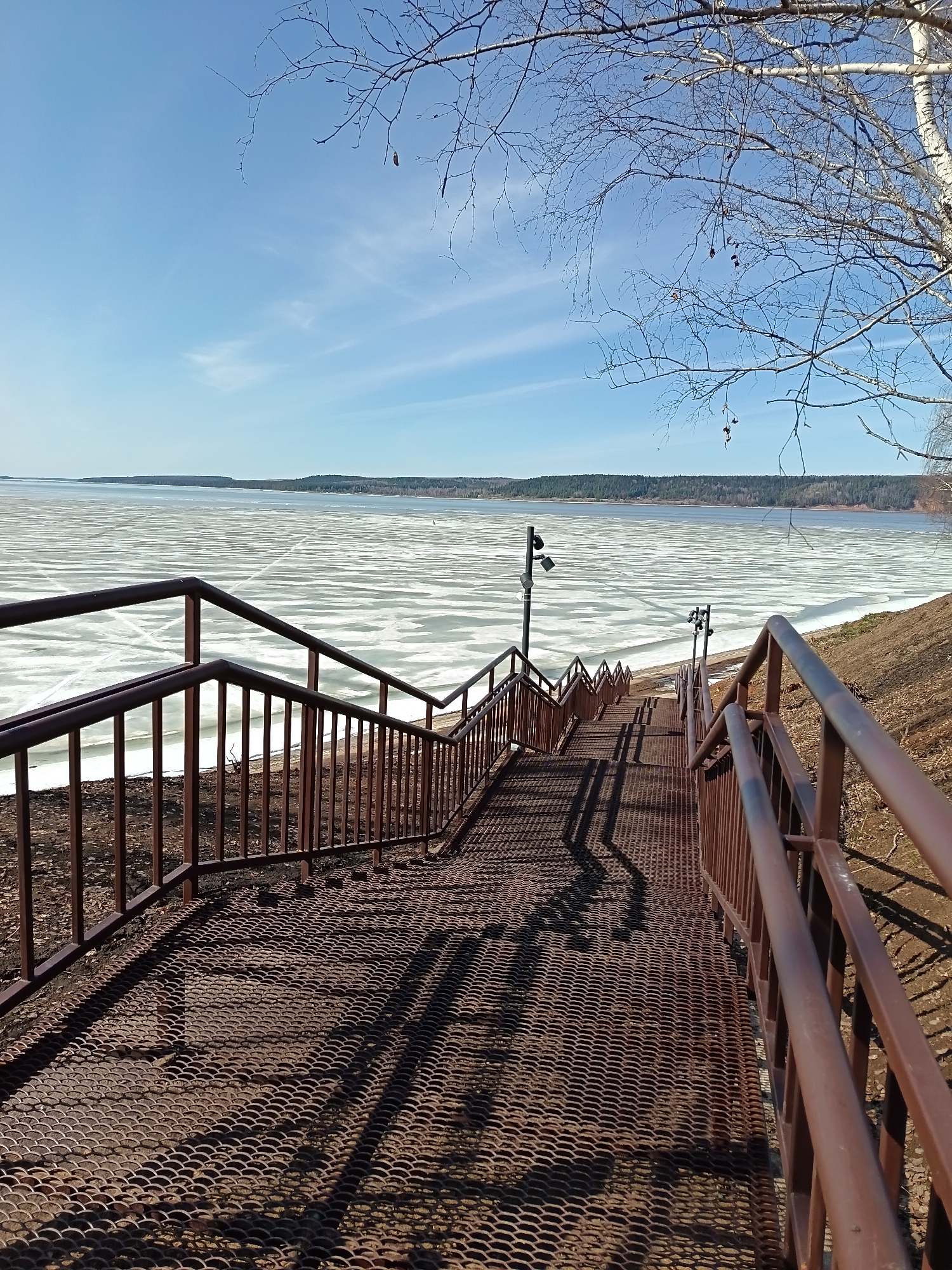An eye-level, outdoor shot presents a downward view of a metal staircase leading towards a partially frozen body of water under a clear blue sky with sparse clouds. The staircase has a perforated metal surface, and brown metal railings are positioned on both sides. Shadows cast by the railings and the staircase structure add depth and texture to the image. In the background, the frozen water extends to the horizon, where a forested shoreline is visible. A few light poles with multiple lights are located along the staircase. A bare tree branch is visible in the upper right corner of the frame.
This is Osa, Russia.