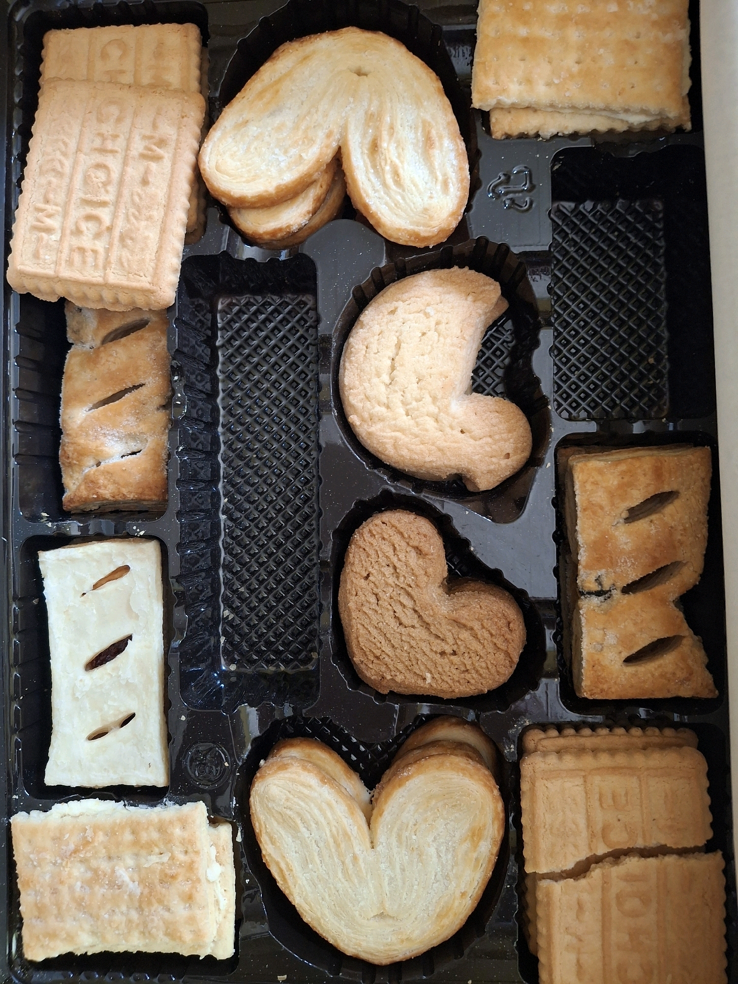 A box of assorted cookies is photographed from above in Lumajang, Indonesia. The cookies are arranged in a black plastic tray. From top left, clockwise: three rectangular cookies with embossed patterns, two palmiers, a stack of two rectangular cookies with a cracked surface, two rectangular cookies with diagonal slits, a heart-shaped cookie, a crescent-shaped cookie, and a rectangular cookie with a slit. The box is partially visible at the top and right edges of the image.