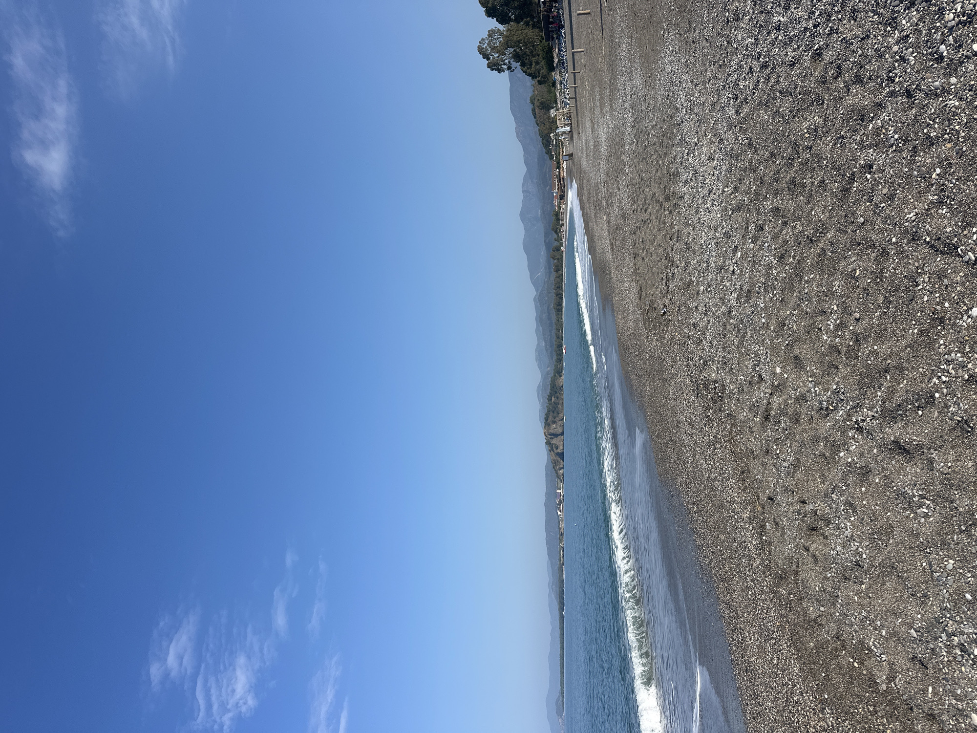 The image depicts a pebbled beach in Fethiye, Turkey, bordered by the Mediterranean Sea on one side and a coastal road and trees on the other. Gentle waves lap the shoreline. In the background, the mountains rise. The sky is predominantly clear and blue with a few wispy clouds.