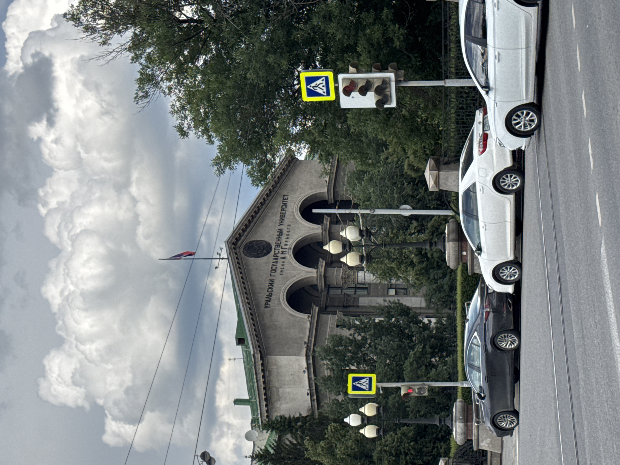 The image depicts the Ural Federal University building in Yekaterinburg, Russia, under a cloudy sky. In the foreground, a dark-colored sedan and two white sedans are parked on a city street. A pedestrian crossing sign and traffic lights are visible on the right side of the image. The university building, characterized by its classical architecture, stands prominently in the background, with trees and overhead wires surrounding it.