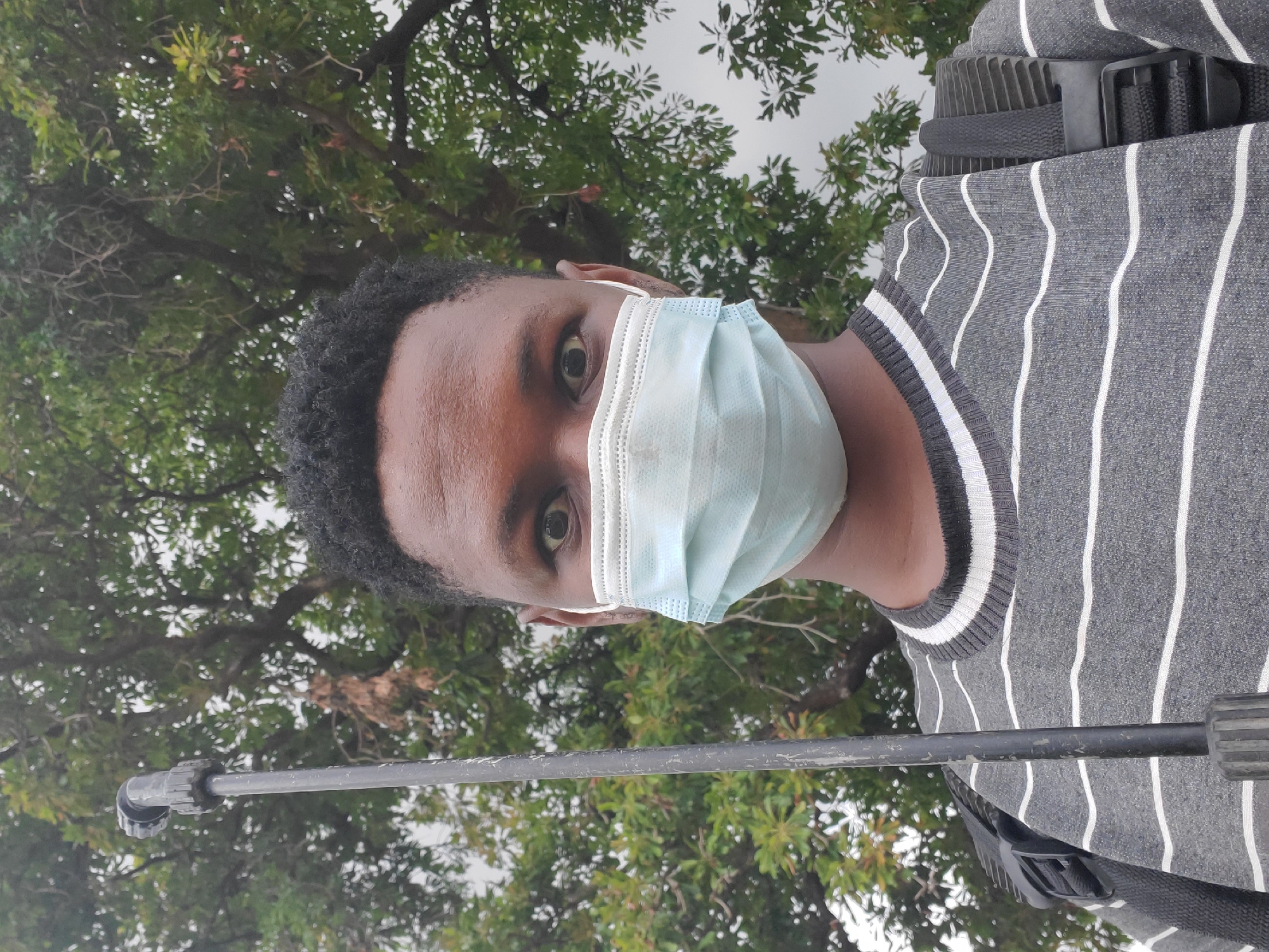A person wearing a light blue surgical mask and a gray and white striped shirt is visible. A black pole is in the foreground. The background features a green tree. The image appears to have been taken outdoors in daylight. Mararaba, Nigeria, is not apparent from the image.