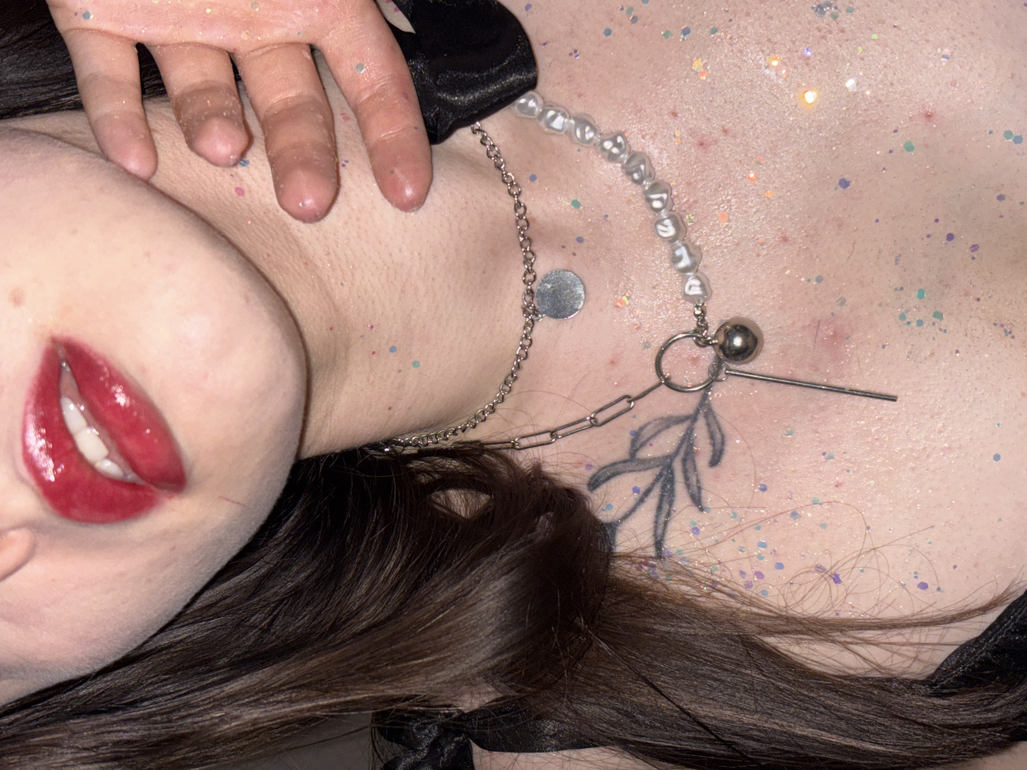 The image depicts a close-up of a person lying down. Their head is tilted, exposing their face, neck, and upper chest. The person has red lipstick and brown hair. They are wearing a pearl necklace, a silver chain with a round pendant, and a choker. A floral tattoo is visible on their chest. The scene includes glitter scattered across the person's skin. There are no discernible features indicating the city of Perm, Russia.