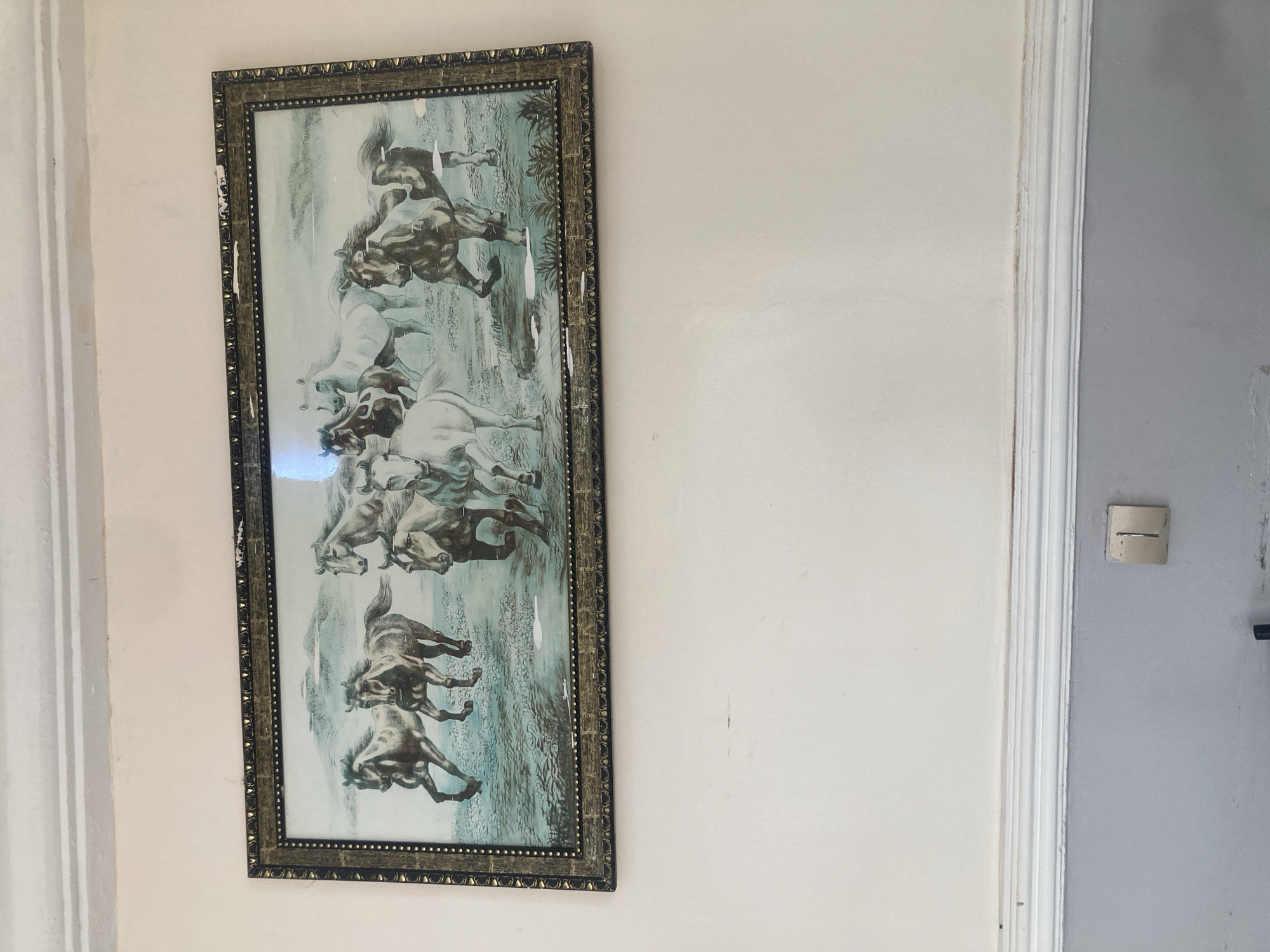 The image shows a framed print of horses hanging on a white wall next to a grey door. The frame is ornate and has some damage. The location is Gura Top, Nigeria.