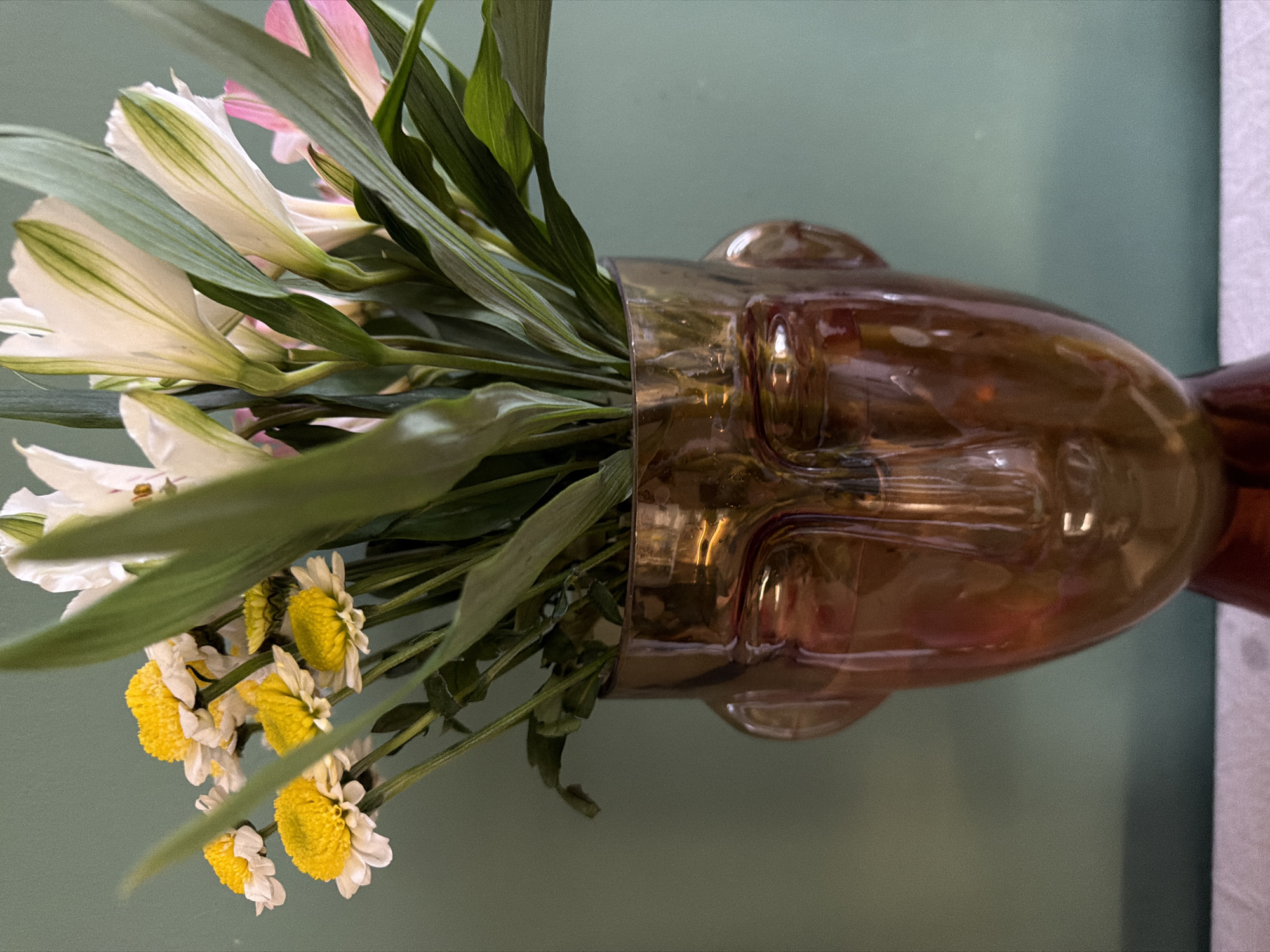 A glass vase shaped like a face contains a floral arrangement featuring white and pink alstroemeria, yellow and white daisies, and green foliage. The vase is translucent amber, revealing the facial features. The background is a muted green wall. The image appears to be taken indoors in the city of Большая Шильна, Russia.