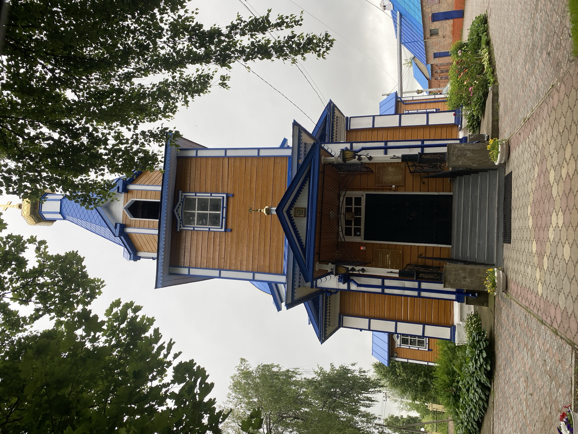 The image shows a wooden Orthodox church with a blue roof and a golden dome, located in the city of Zelenodol'sk, Russia. The church is painted in warm brown tones with white and blue trim. It has a prominent entrance with a covered porch and stairs leading up to the double doors. The church is surrounded by greenery, including trees and bushes, and a paved walkway leads to the entrance. In the background, other buildings with blue roofs are visible. The sky is overcast. There are no people or animals in the image.