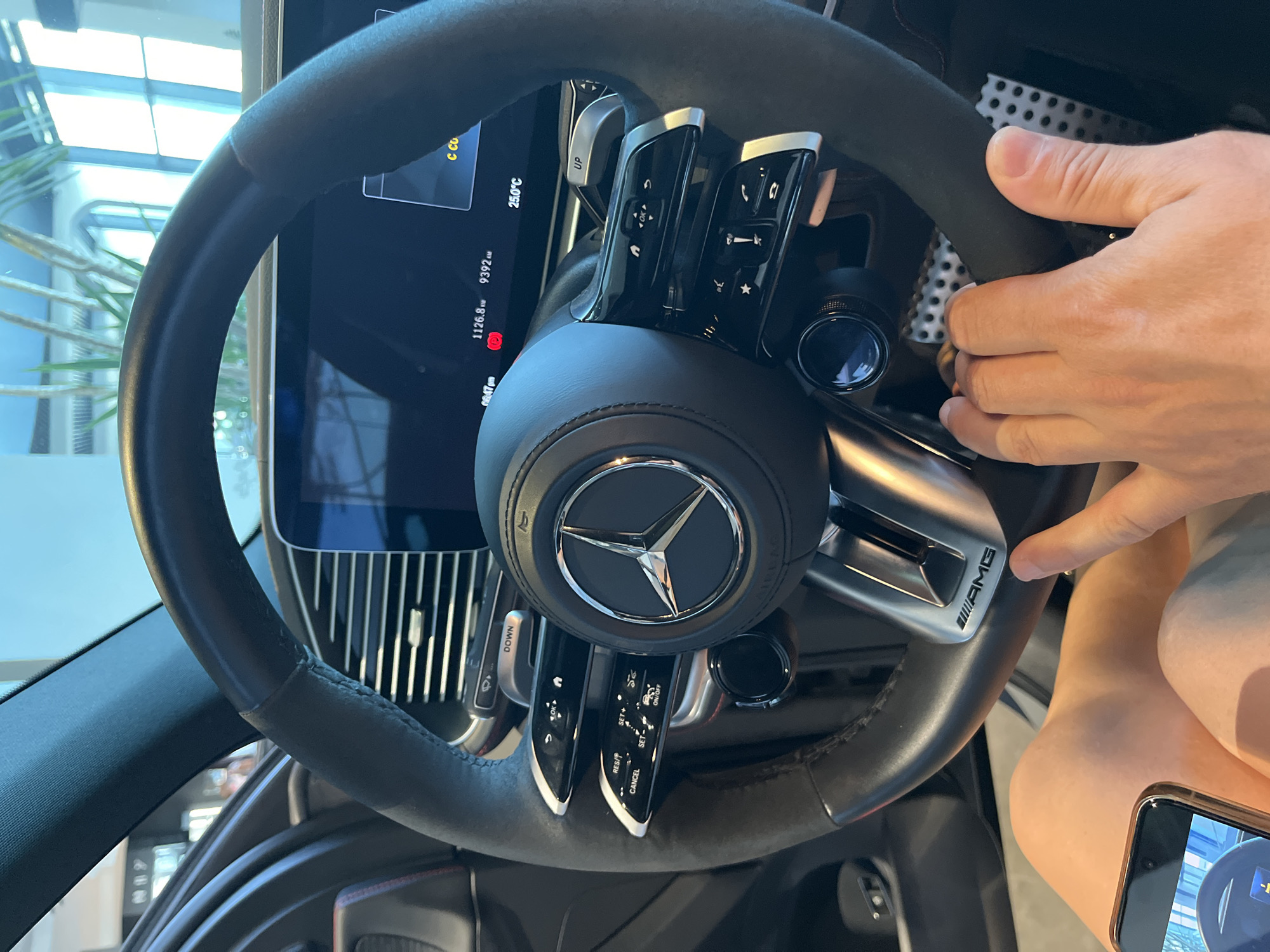 A close-up shot captures a person's hands on the steering wheel of a Mercedes-Benz AMG car. The steering wheel is black leather with silver accents, and the Mercedes-Benz logo is prominently displayed in the center. The digital dashboard is visible in the background, showing vehicle information. The person's legs are also visible, clad in light-colored shorts. The scene appears to be indoors, possibly in a car dealership or showroom. The image does not provide any information about the city of Sochi, Russia.