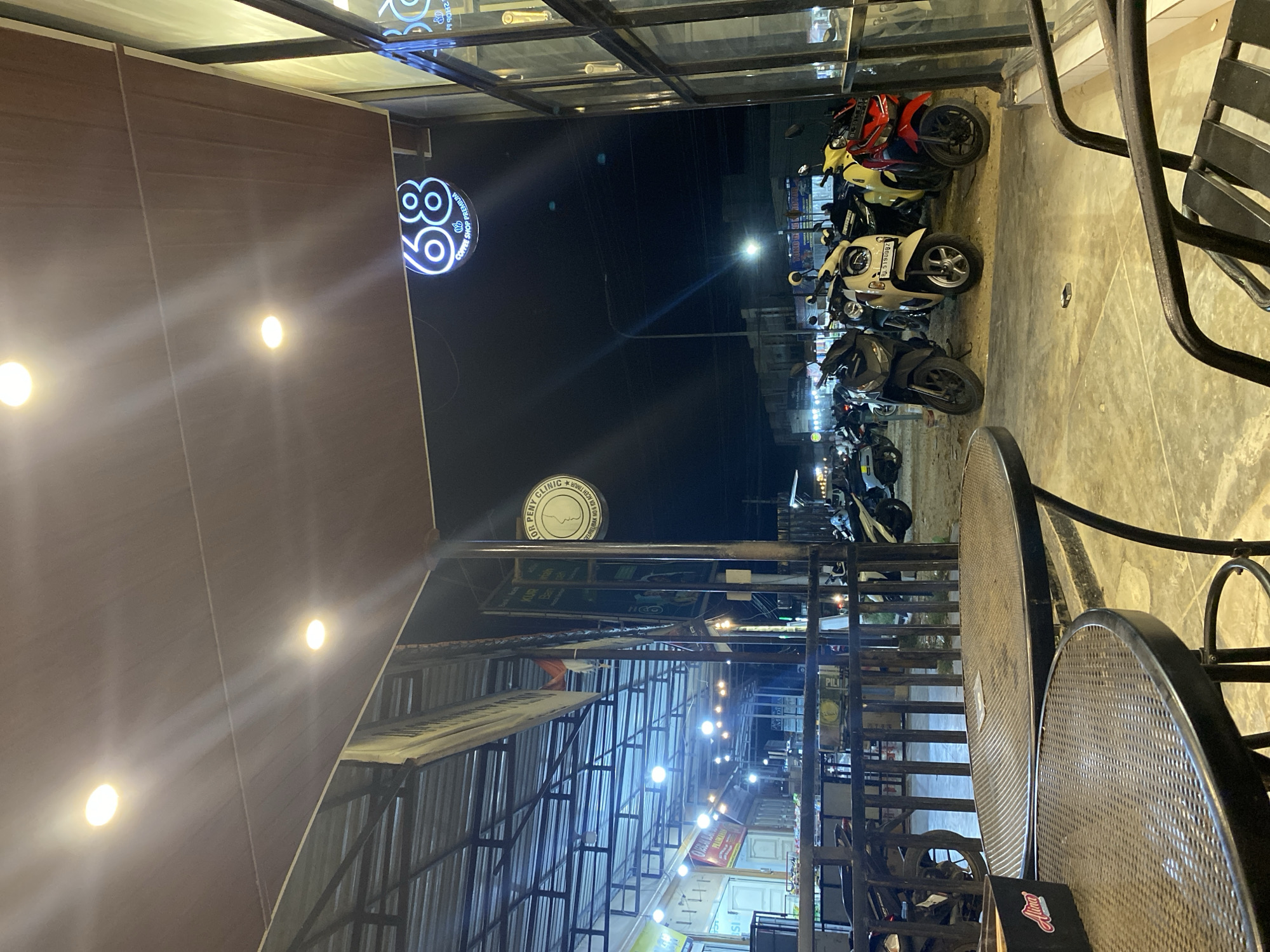 Here is a description of the image:
The image depicts a street scene in Tanjongkapai, Indonesia at night. Several parked motorbikes are visible along the street's edge. Overhead, a series of lights illuminate the area, while a sign for "68 Coffee Shop Premium" and "DR PENY CLINIC WILHE" is prominent. Other shops and businesses are visible in the background, with various signs indicating different establishments. Tables and chairs, seemingly part of an outdoor seating area, are positioned to the right side of the frame.