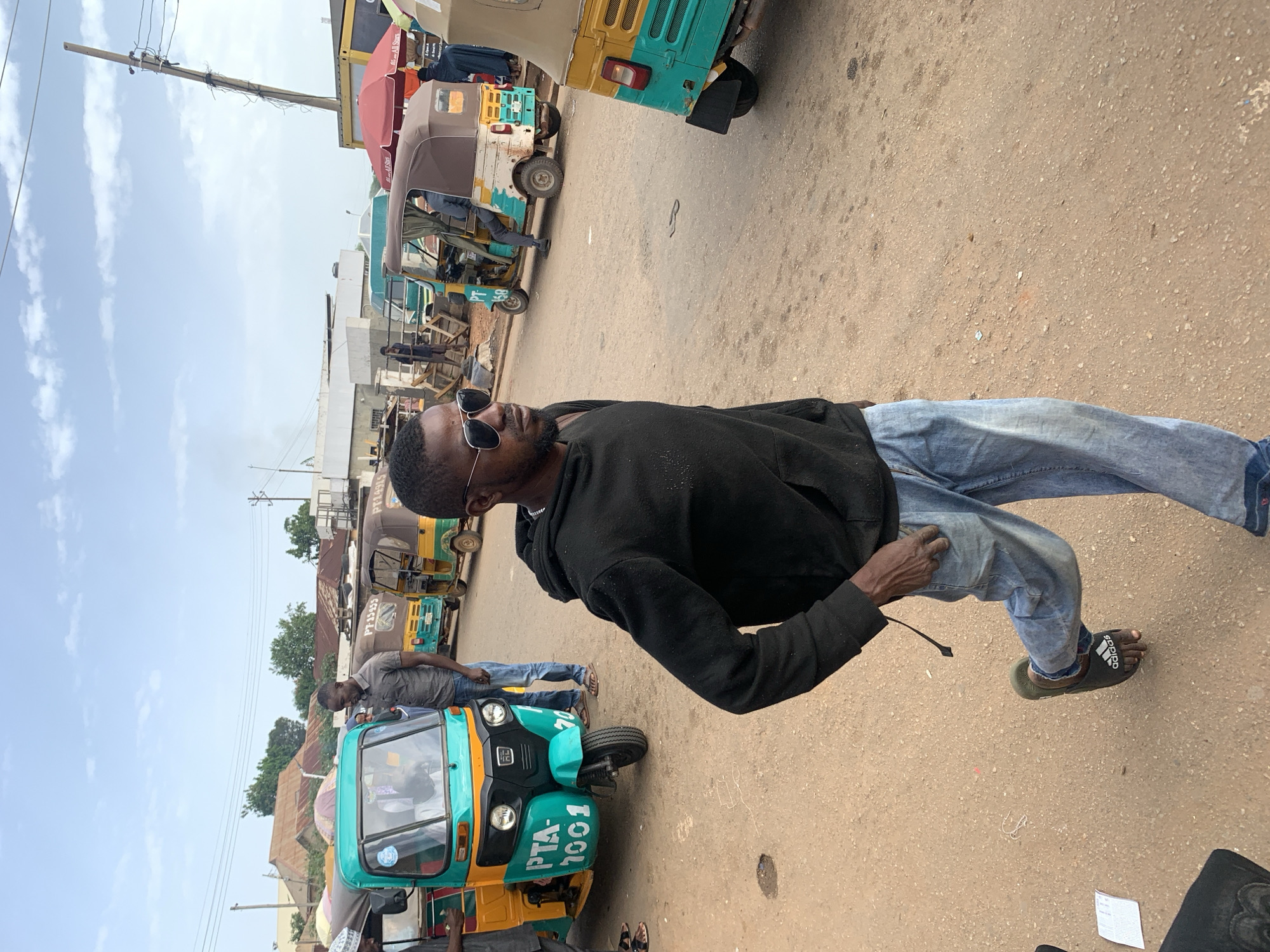 A man wearing sunglasses and a black hoodie walks on a dirt road in Gura Top, Nigeria. He is wearing light blue jeans and dark green Adidas slides. In the background, several three-wheeled vehicles, commonly known as "Keke NAPEPs," are parked. Some are teal and yellow, while others are brown and yellow. The road is unpaved, with scattered debris. Buildings with corrugated metal roofs are visible in the distance, along with utility poles and wires. The sky is partly cloudy.