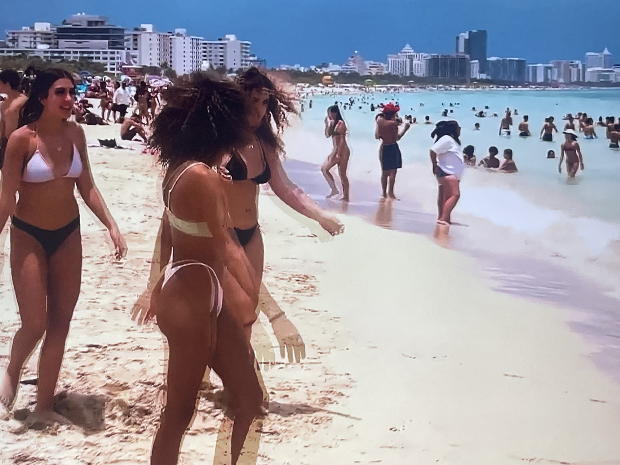 This media file captures a vibrant beach scene. The subjects are a group of people, predominantly women in bikinis, enjoying a sunny day. The setting is a sandy beach with the ocean stretching out to the horizon. In the background, a cityscape with modern buildings is visible, hinting at an urban coastal location.
The activity taking place is leisure and recreation. People are seen walking on the sand, wading in the shallow water, and swimming in the ocean. The mood appears relaxed and cheerful, with individuals socializing and enjoying the summer weather.
Notable details include the bright sunlight, suggesting it's daytime, likely late morning or afternoon. The clear blue sky indicates good weather conditions. The architecture of the background buildings provides a clue to the location, but given the lack of specific Russian architectural elements, and the typical appearance of a crowded beach, it is unlikely to be Shakhovskaya, Russia. There is no visible text in the image.