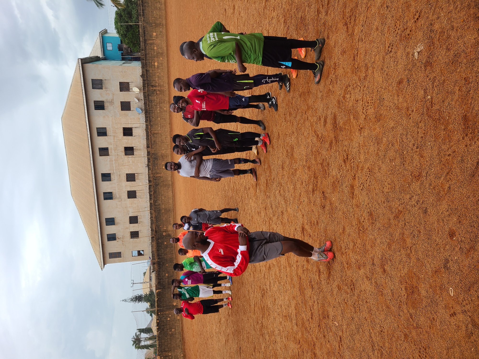 A wide, low-angle shot depicts a group of approximately a dozen men standing on a dirt field, possibly engaging in a training exercise. They are lined up facing the camera, with their bodies oriented towards the right side of the frame.
The men are wearing various athletic attire, including jerseys in red, green, purple, white, and black. The man closest to the camera is wearing a red and white jersey with grey shorts. The field is an even dirt surface, likely a sports field.
In the background, a large, multi-story building is visible, positioned along the left edge of the frame. The building is light in color with dark windows. Above the building is a cloudy sky.
The image appears to be taken outdoors in a location identified as Unknown, Unknown. The setting suggests a sports training facility or a recreational field within a city.