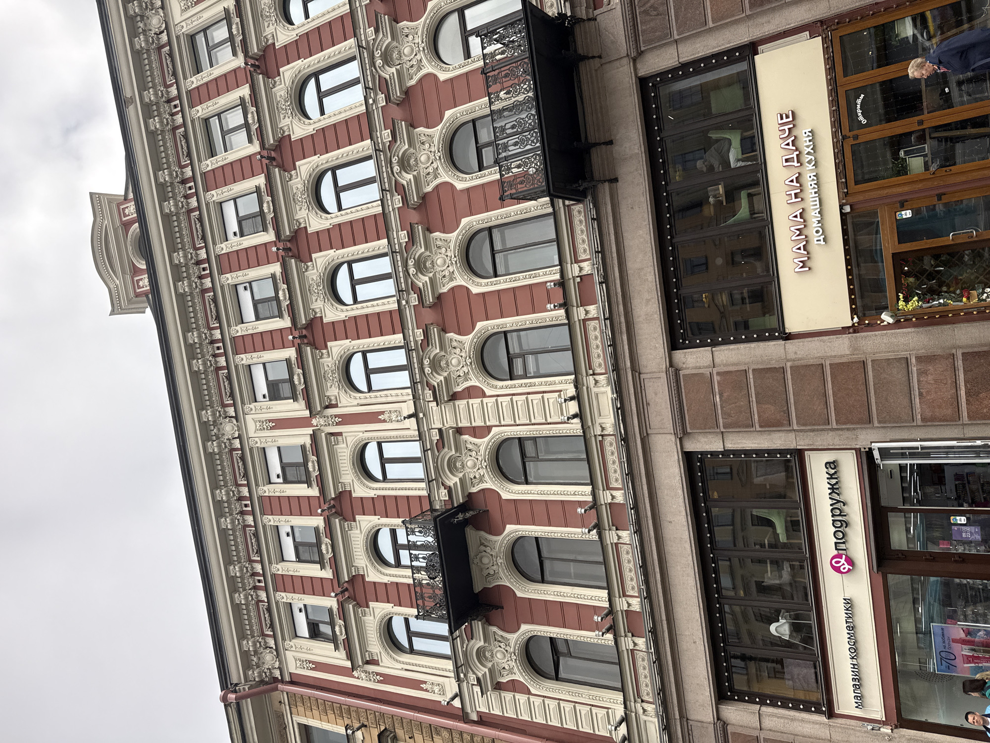 The image shows a building in Saint Petersburg, Russia. The building has a red and white facade with ornate architectural details, including balconies and window frames. On the ground floor, there are two storefronts. One sign reads "MAMA HA ДAЧE ДОМАШНЯЯ КУХНЯ" (Mama on the Dacha Home Cuisine) and the other says " подружка МАГАЗИН КОСМЕТИКИ " (Girlfriend Cosmetics Store).
