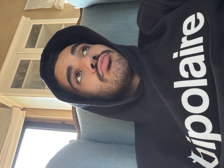 An indoor, close-up photograph captures a single male individual, appearing to be in his 20s or 30s, seated in a light blue upholstered armchair. The individual has a medium-dark complexion, dark hair, brown eyes, and a moustache with beard stubble. He is wearing a black hooded sweatshirt with the hood pulled over his head. The word "bipolaire" is printed in white, stylized capital letters across the chest of the sweatshirt. The individual's head is slightly tilted upwards and to the right, with his eyes gazing off-frame in that direction, and his lips are pursed in a pout. The background shows an interior space featuring a bright window on the left, an adjacent light-colored wall, and a white wooden cabinet with glass doors displaying glassware, positioned above and behind the subject's head. The scene is located indoors in Mauléon-d'Armagnac, France.