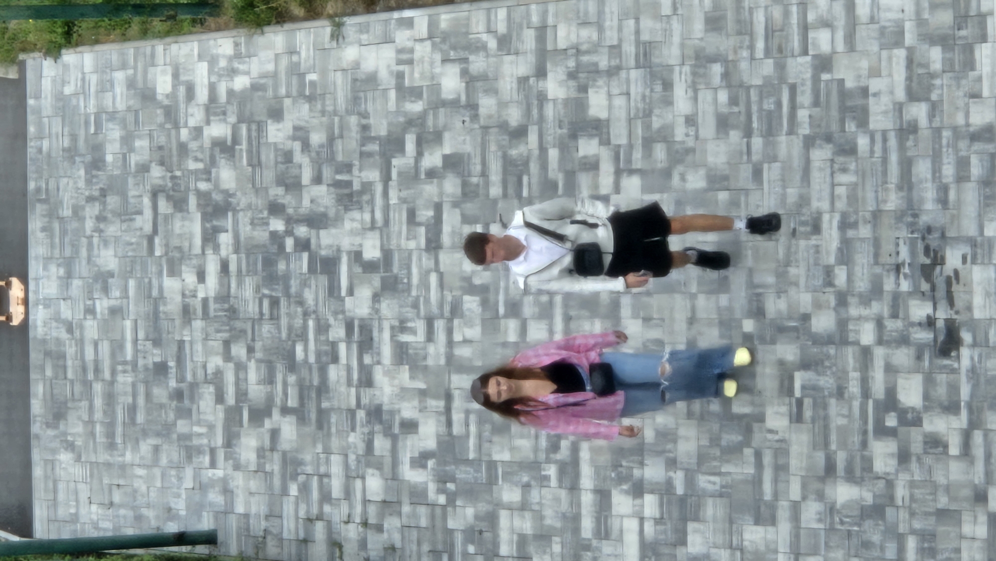 In Prešov, Slovakia, two individuals are captured walking along a textured, grey, rectangular paver pathway. The female, on the left, wears a pink plaid shirt, blue ripped jeans, and yellow shoes, with a black crossbody bag around her waist. The male, on the right, is dressed in a white polo shirt, black shorts, and black sneakers, also carrying a black crossbody bag. The pathway shows signs of wear and tear, with some discoloration and staining. The overall environment appears to be an outdoor public space, possibly a park or promenade, with hints of greenery and fencing visible along the edges.