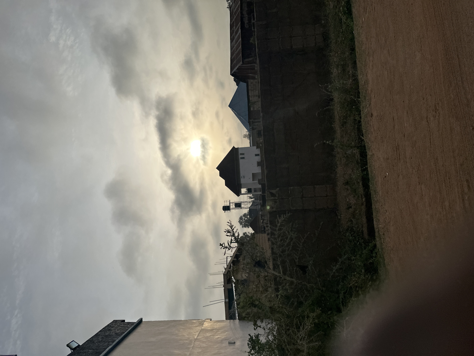 The image depicts an outdoor scene in Bukuru, Nigeria, characterized by overcast skies with the sun partially obscured by clouds. In the middle ground, a residential area with houses and a water tank is visible. To the right, there is a dark, likely overgrown or unkempt area adjacent to a reddish-brown path or road. The foreground features a lower structure, possibly a building roof, partially illuminated by the ambient light. No people are visible in the frame.