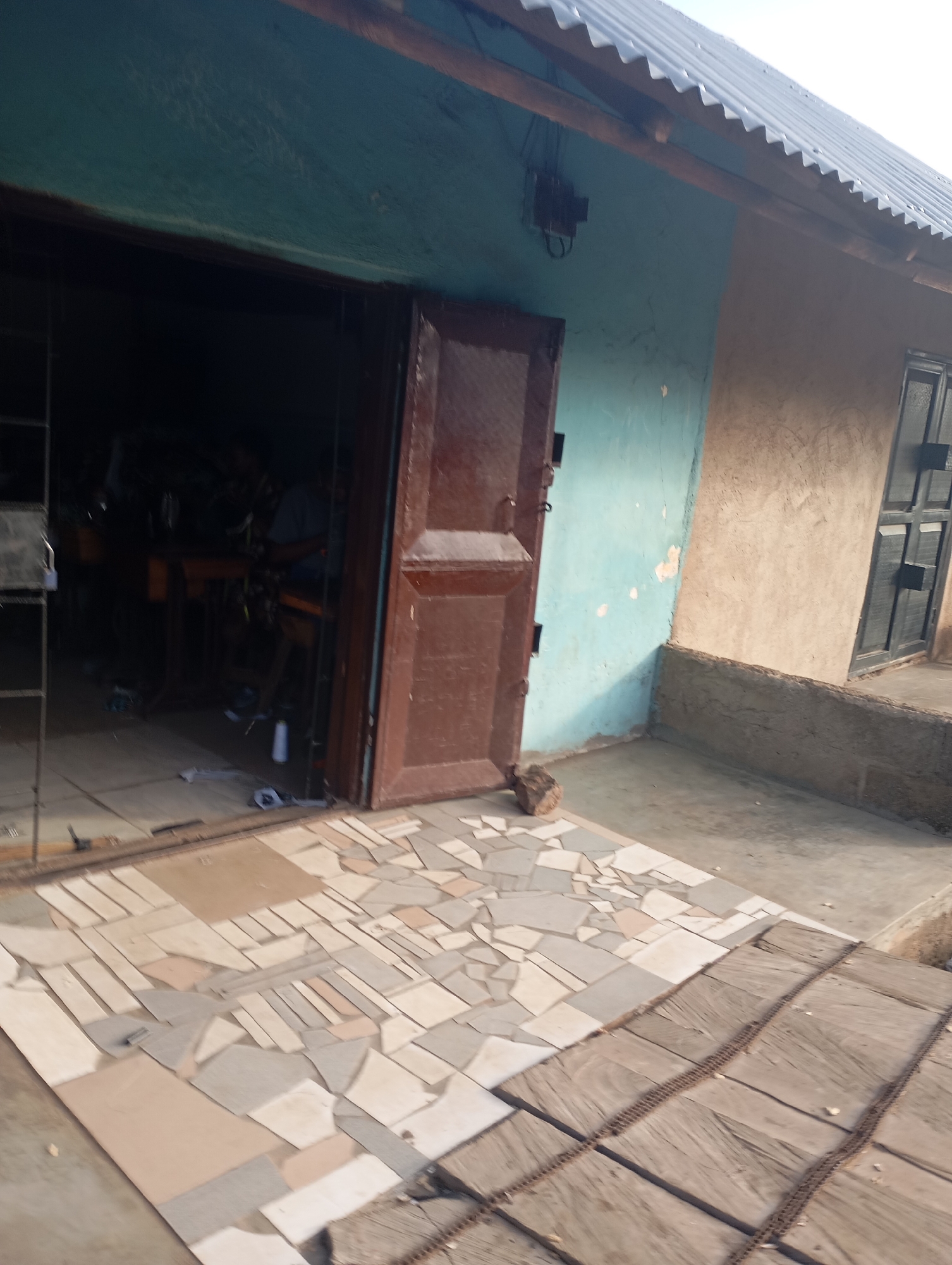 The image shows the entrance to a workshop in Shendam, Nigeria. The workshop appears to be a tailor's shop, with sewing machines visible inside. The exterior of the building is a mix of blue and tan plaster. The entrance is a large, wooden double door, one of which is ajar. A mosaic tile path leads to the entrance. The roof is corrugated iron. The scene is weathered and shows signs of use.