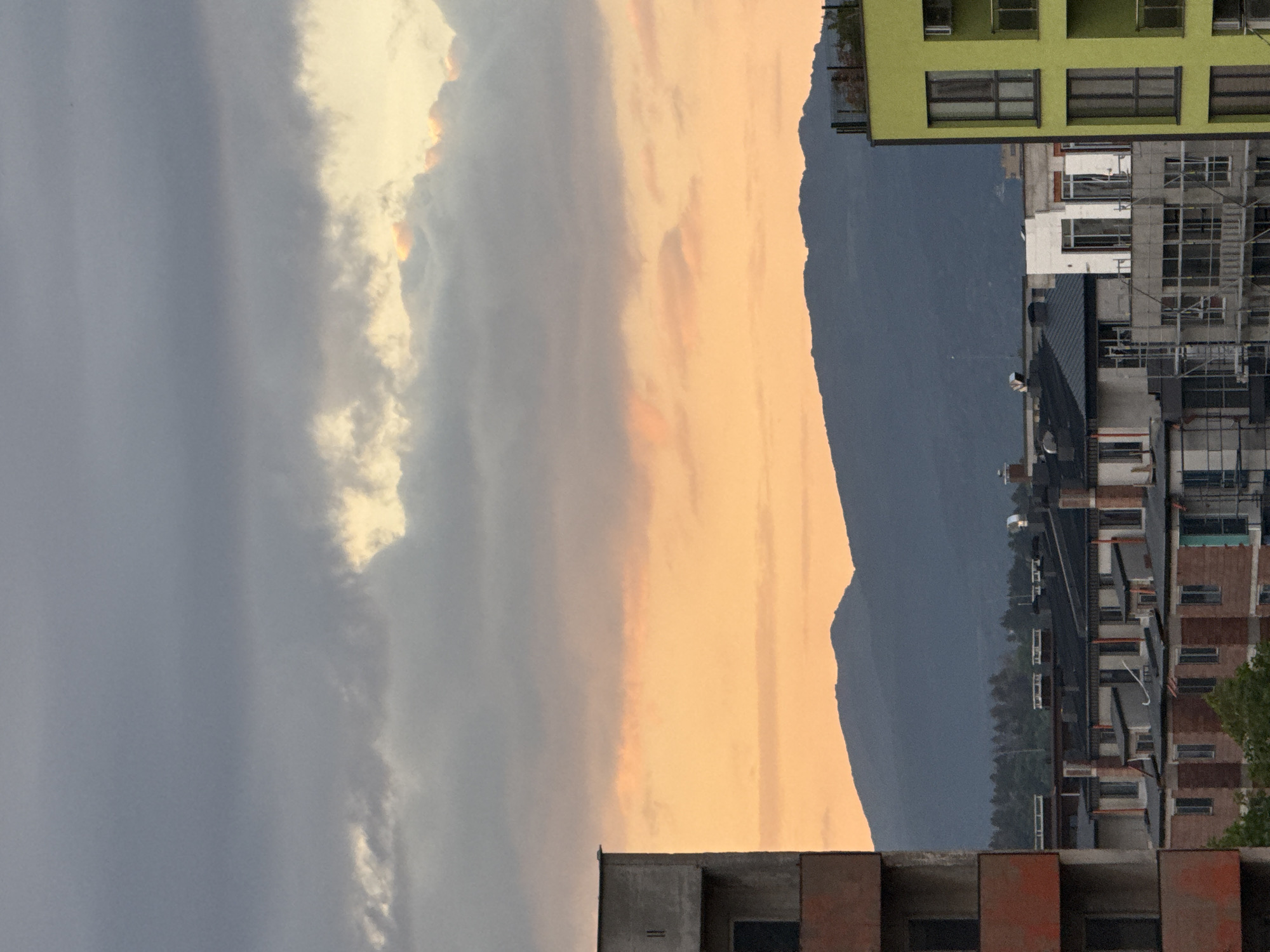 The image captures a partially obstructed view of the Vitosha Mountain in Sofia, Bulgaria, likely taken from a high vantage point within the city. To the left, a large cloud formation dominates the sky, transitioning from a dark grey to a lighter, sunlit white. A band of orange sky separates the clouds from the mountain range. The mountain appears as a dark silhouette against the bright sky.
In the foreground, sections of buildings are visible. On the right, there is a green building with multiple windows, next to a building under construction, with scaffolding visible. Below, another building with visible balconies can be seen. In the bottom center, there is an unfinished rooftop with an exposed brick layer.