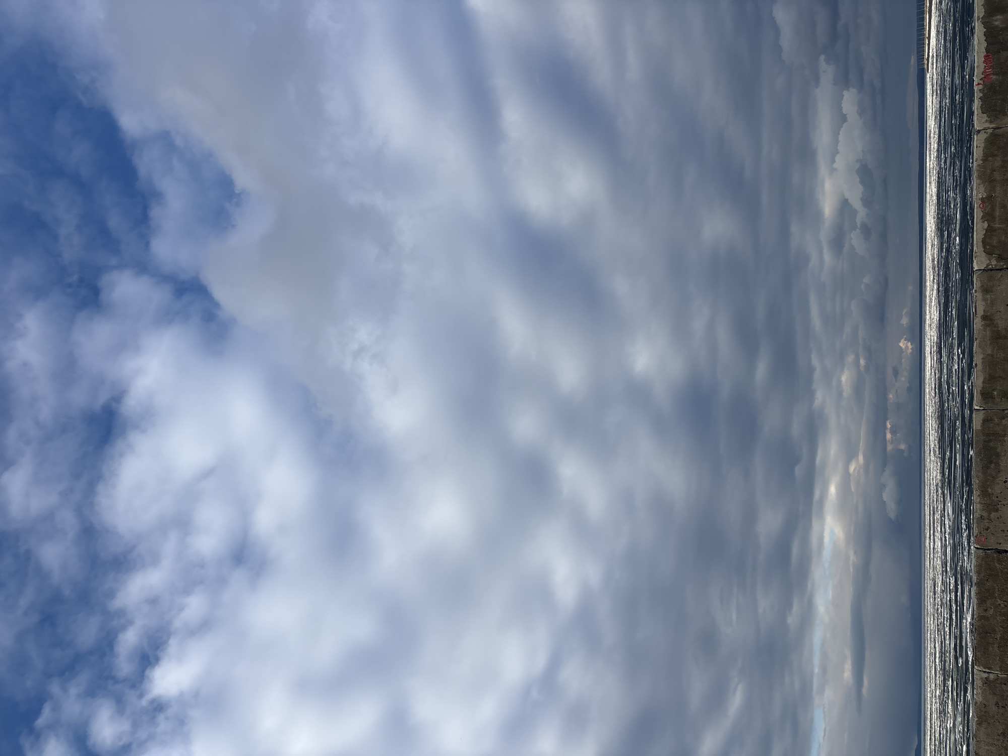 The image presents a broad outdoor scene, dominated by a partially cloudy sky and a body of water, with a concrete structure marking the foreground.
The sky, occupying the majority of the frame, transitions from a clear, bright blue with scattered white, wispy clouds on the left to a more dramatic expanse of dense, layered grey clouds that stretch horizontally across the center and right. Sunlight intermittently penetrates these clouds, creating brighter patches and highlighting their textured forms.
Below the cloudscape, a large body of water, likely a river or reservoir near Chaikovsky, Russia, extends towards a distant horizon. Its surface reflects the overhead light, creating shimmering, vertical streaks that indicate ripples or slight waves under the mixed sunlight and cloud cover.
Along the far right and bottom edge of the image, a robust, weathered concrete wall or embankment is visible. This dark grey structure has a rough texture, showing signs of exposure to the elements. Faint, red, indecipherable markings or smudges are present on its surface.
The scene depicts a peaceful, natural landscape. The time of day appears to be daytime, with a play of light and shadow suggesting either morning or late afternoon sun breaking through varying cloud densities. No people, specific activities, or legible text are visible.